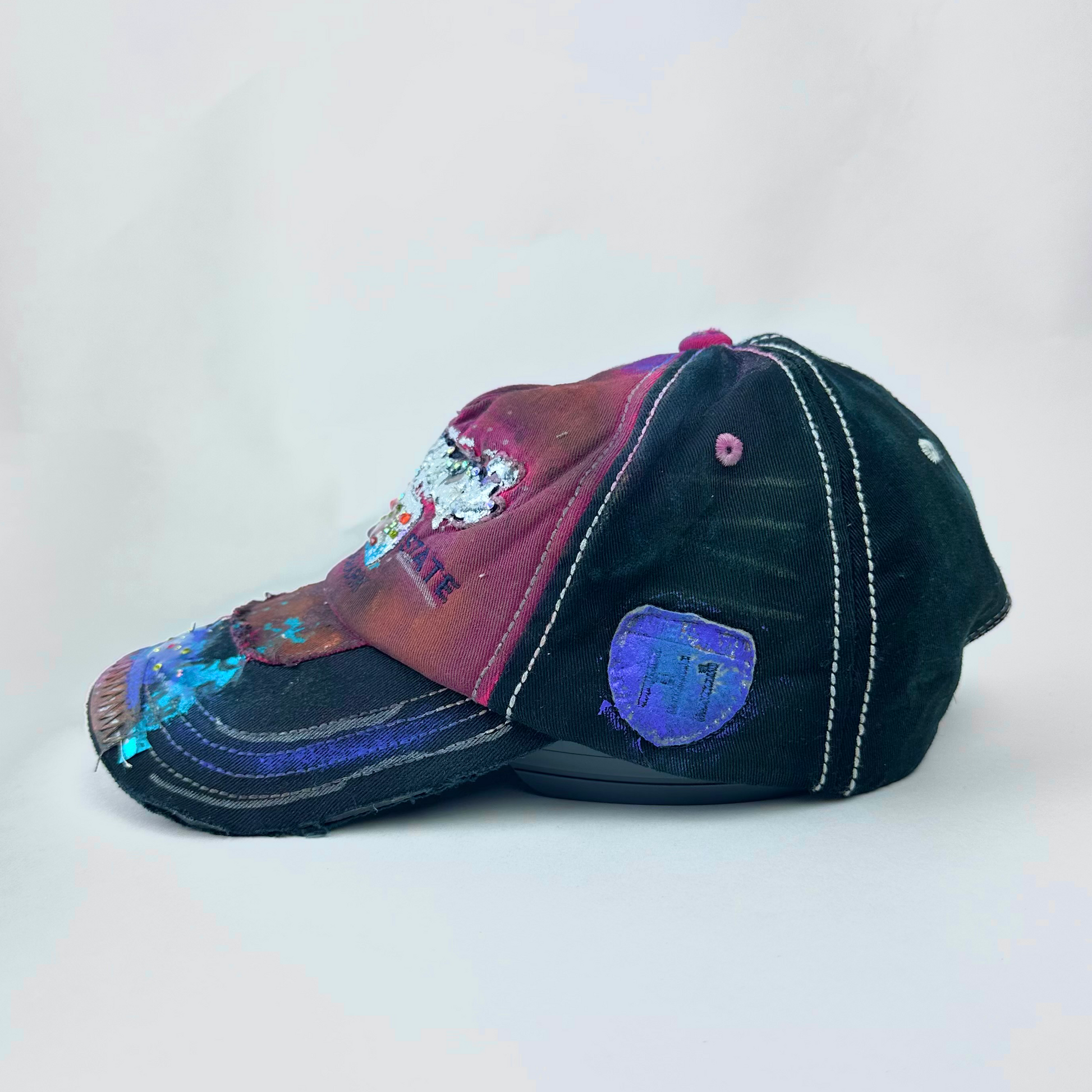 ASTROBOI* 'Hibiscus' red and black Y2K Hawaii rhinestone hat with silver embroidered 'Hawaii ALOHA STATE TRADEMARK' text, purple and pink paint splatter, and metallic rhinestone embellishments, right side.