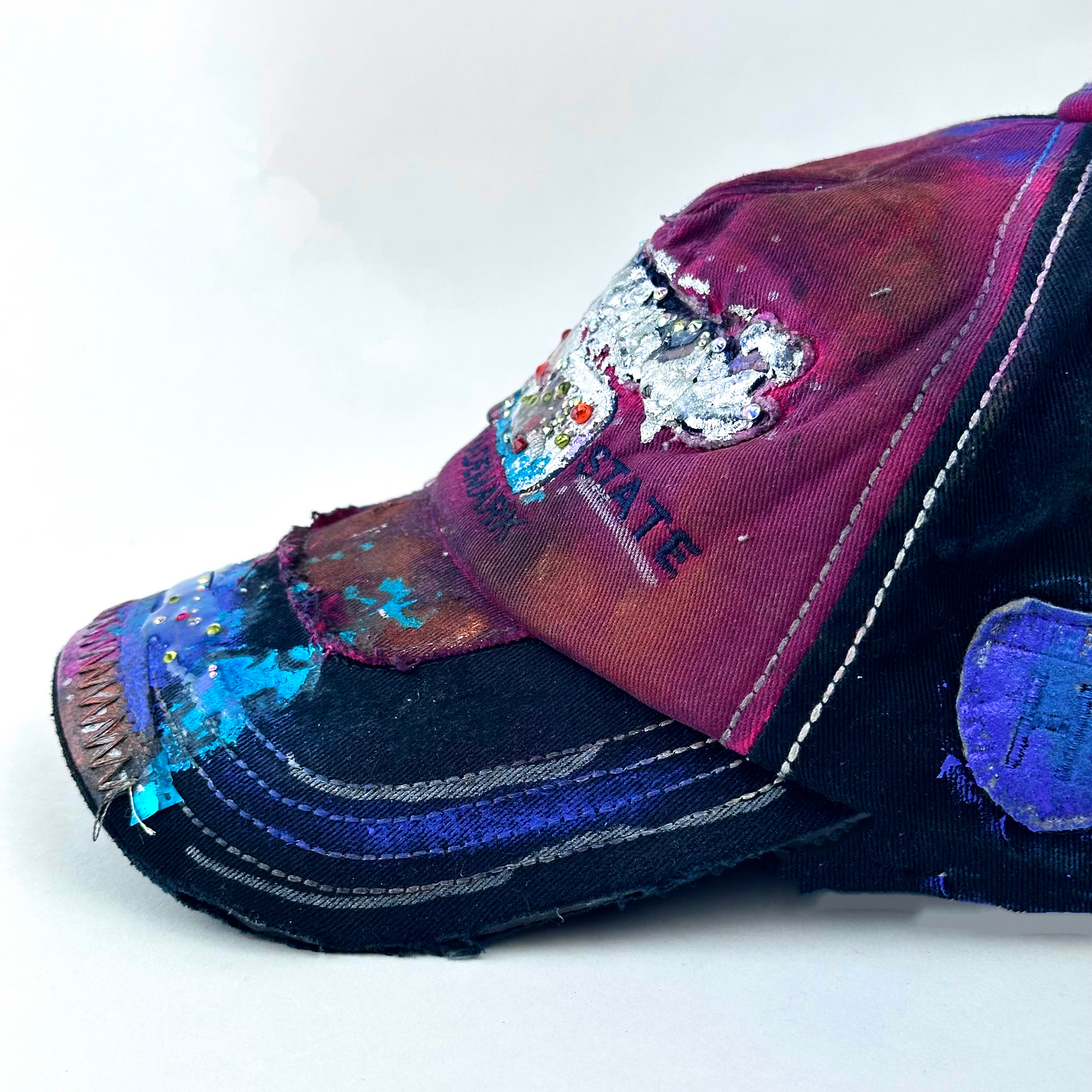 ASTROBOI* 'Tropicale' red and black Y2K Hawaii rhinestone hat with silver embroidered 'Hawaii ALOHA STATE TRADEMARK' text, purple and pink paint splatter, and metallic rhinestone embellishments, right side visor detail.