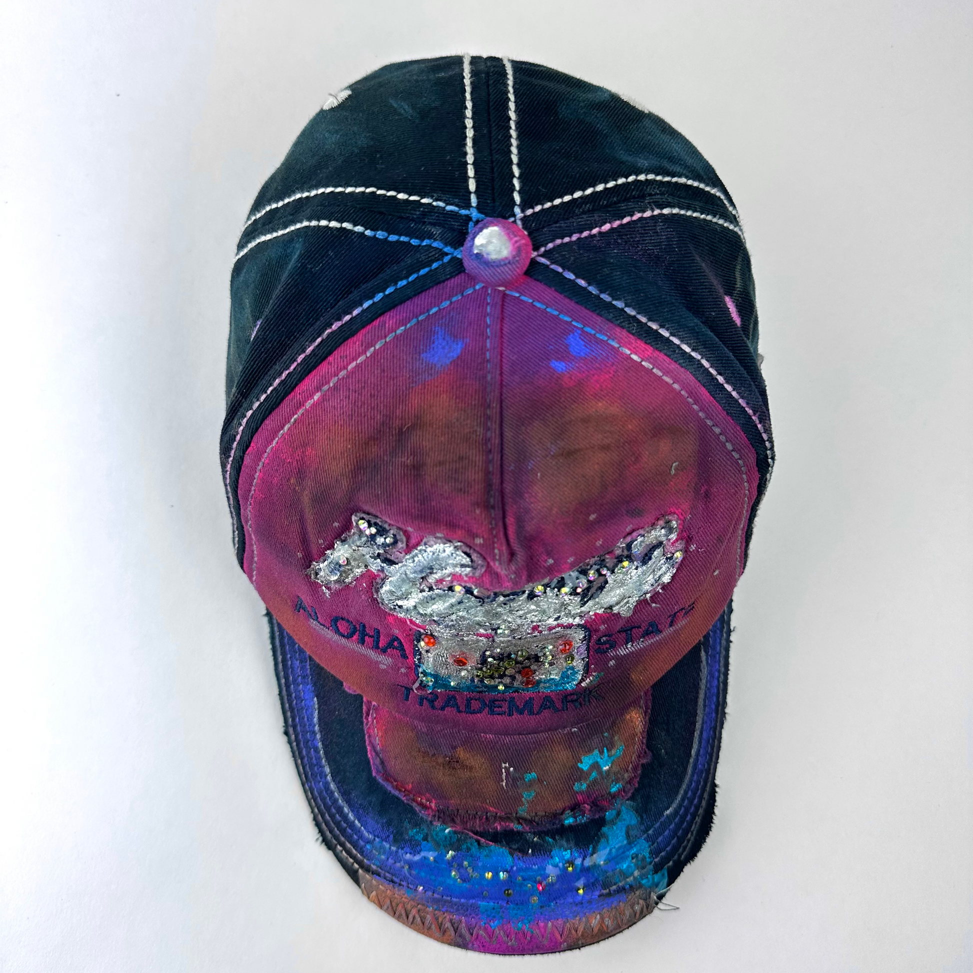 ASTROBOI* 'Hibiscus' red and black Y2K Hawaii rhinestone hat with silver embroidered 'Hawaii ALOHA STATE TRADEMARK' text, purple and pink paint splatter, and metallic rhinestone embellishments, top view.