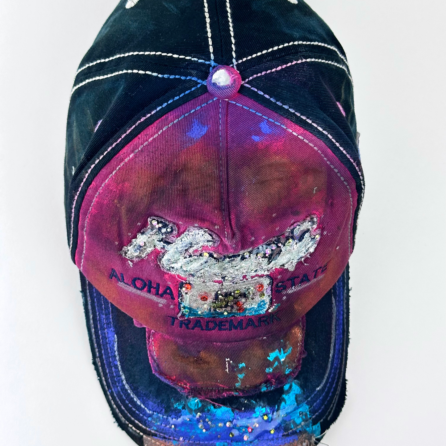 ASTROBOI* 'Hibiscus' red and black Y2K Hawaii rhinestone hat with silver embroidered 'Hawaii ALOHA STATE TRADEMARK' text, purple and pink paint splatter, and metallic rhinestone embellishments, top view.