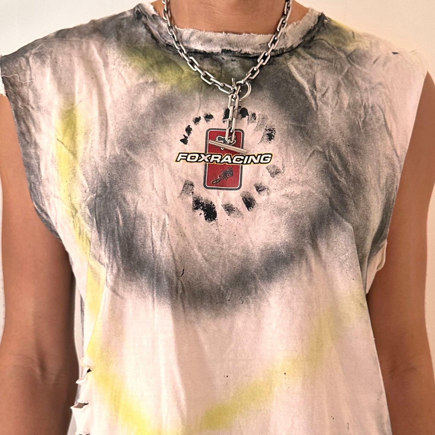 Close-up of ASTROBOI* x Fox Racing tank showing black and acid yellow airbrush streak detailing, punched holes, and raw hem finish on white fabric.