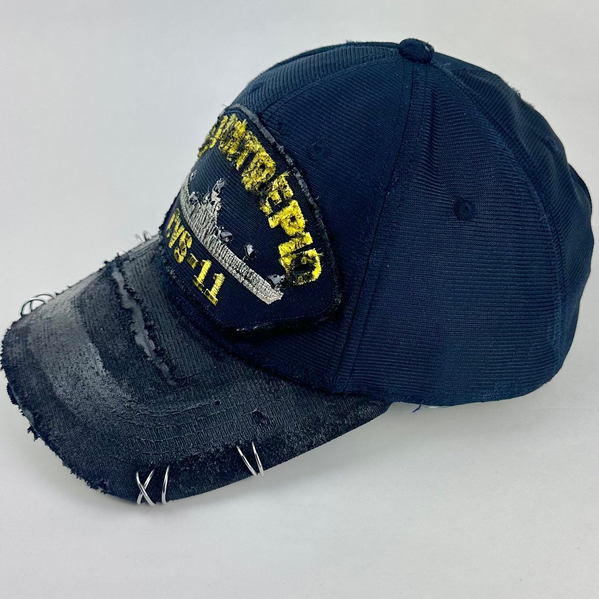 Left side of ASTROBOI* upcycled Navy blue and black USS Intrepid CVS-11 snapback hat with silver rhinestone-embellished front patch, stainless steel brim piercings, and hand-painted black visor on a neutral background.