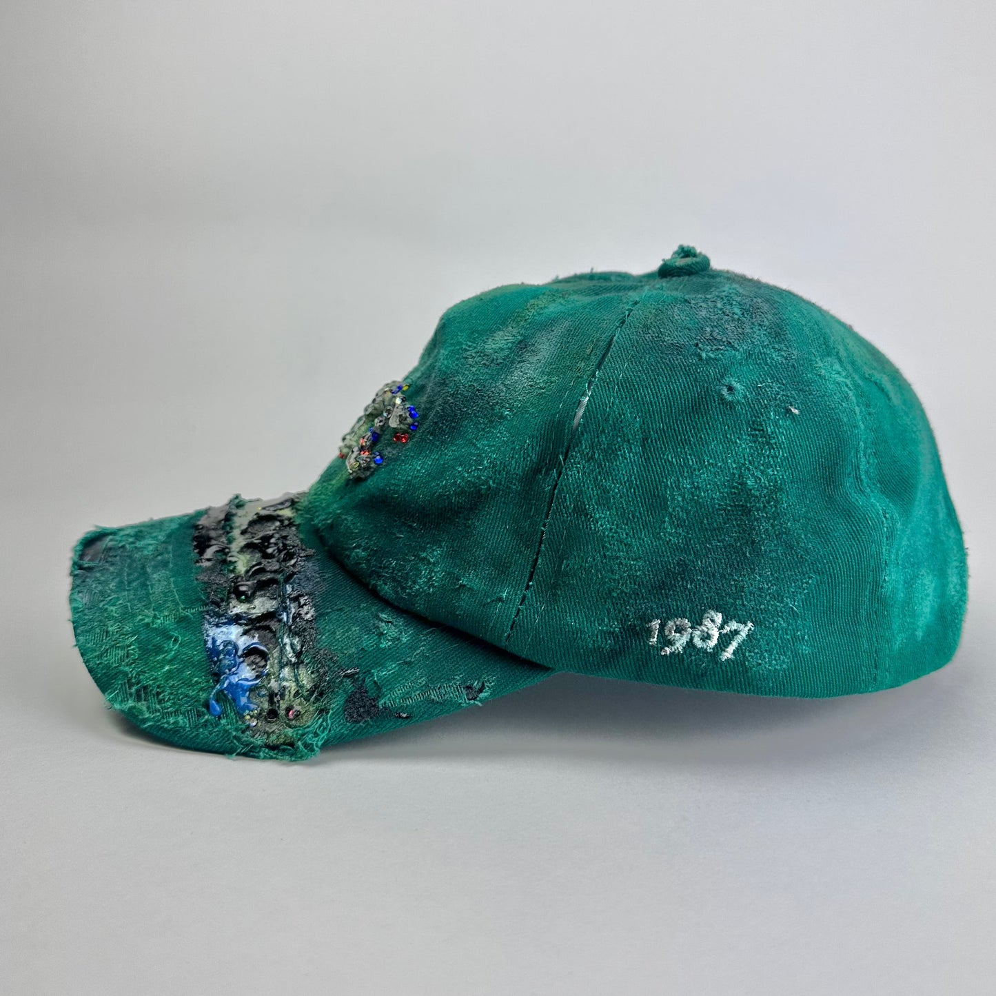 Left side of ASTROBOI* Smith NYC green distressed hat with clear rhinestone logo, custom paint treatment and blue black resin textured visor.