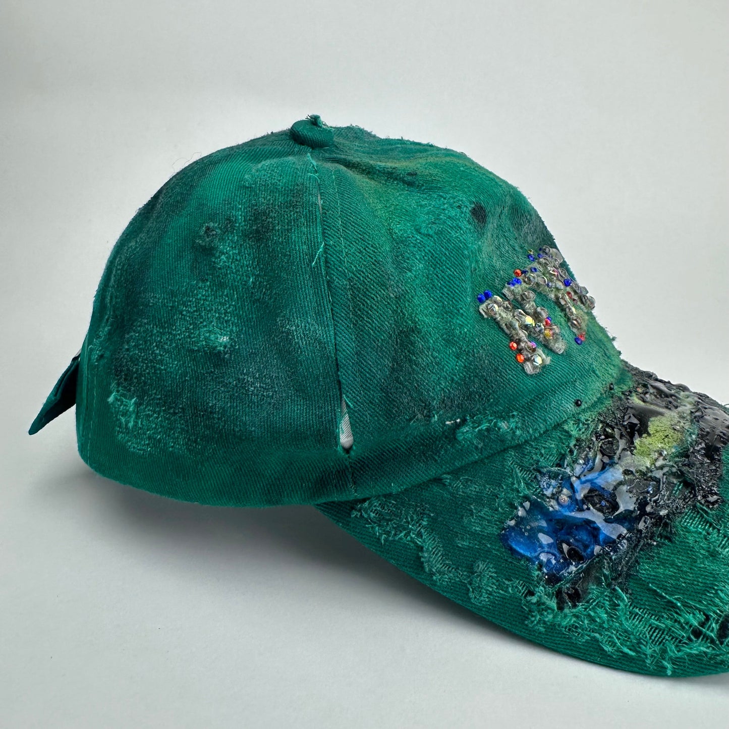 Right side detail on ASTROBOI* Smith NYC green distressed hat with clear rhinestone logo, custom paint treatment and blue black resin textured visor.