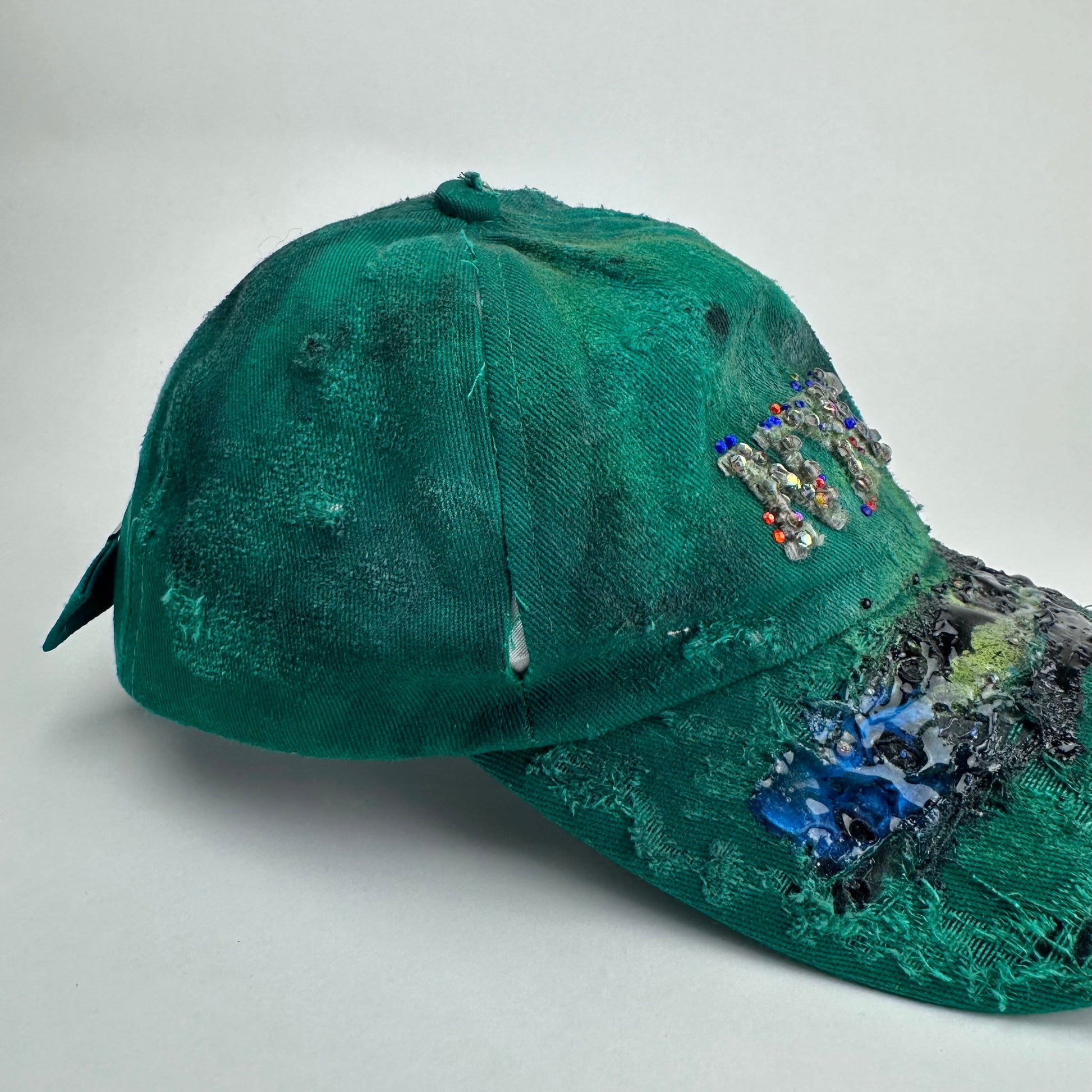 Right side detail on ASTROBOI* Smith NYC green distressed hat with clear rhinestone logo, custom paint treatment and blue black resin textured visor.