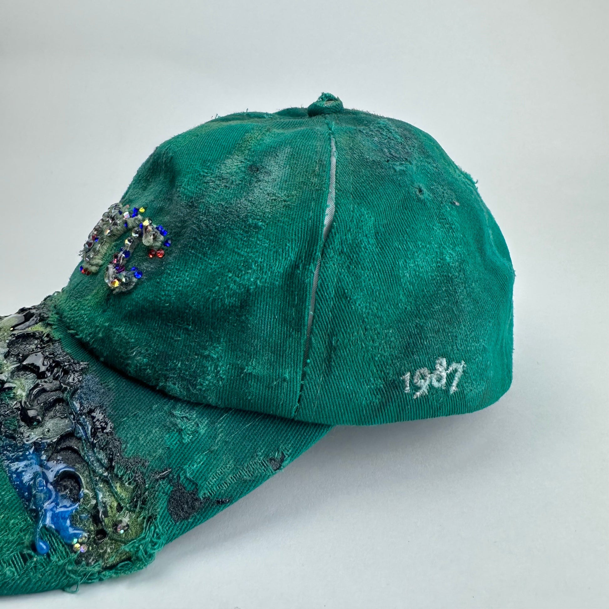Left side open seam detail on ASTROBOI* Smith NYC green distressed hat with clear rhinestone logo, custom paint treatment and blue black resin textured visor.