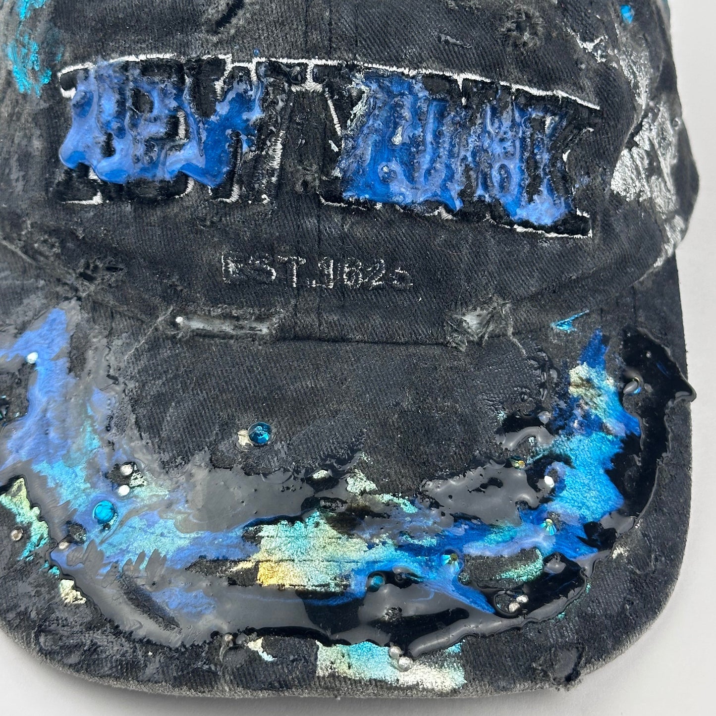 Close up detail of ASTROBOI* Plavala black iridescent hat with rhinestone NYC logos, metallic paint and black clear resin creating a hazy milky way effect.