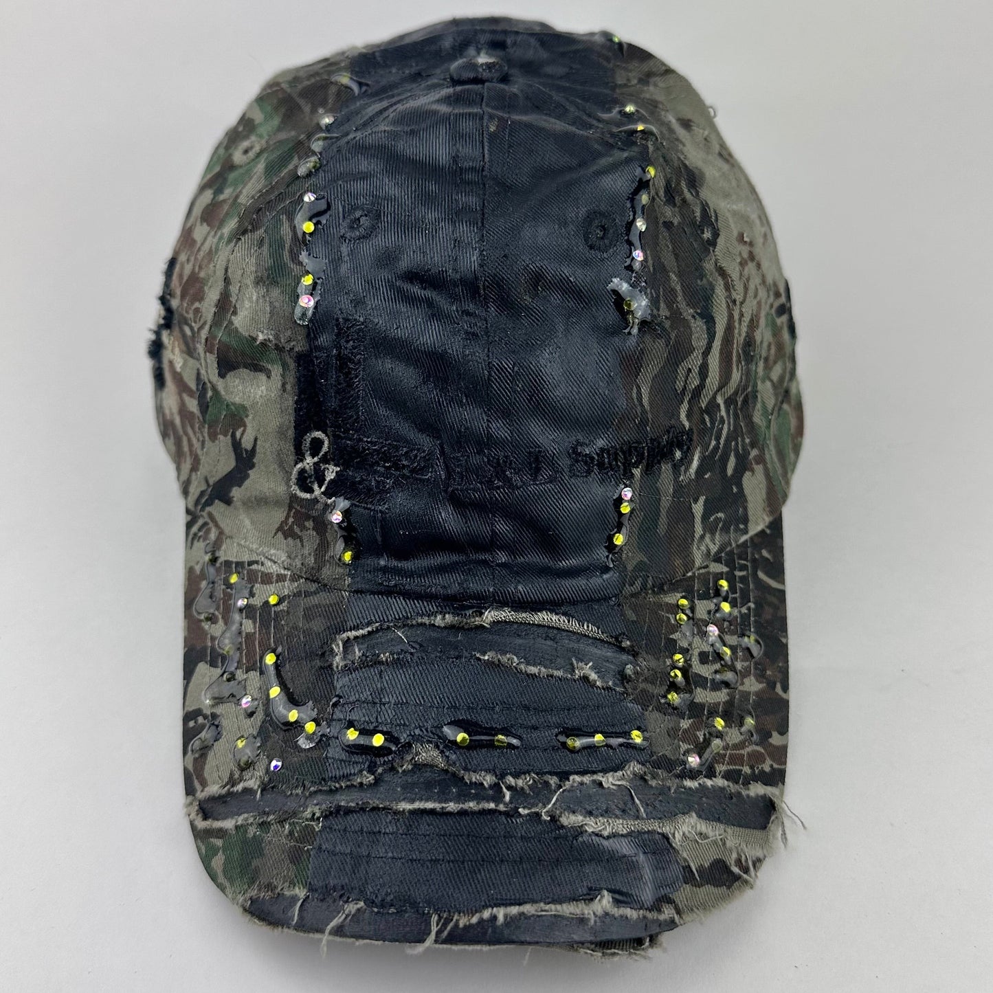 Front flat lay of ASTROBOI* L&L Supply green RealTree camo utility hat with purple and green rhinestones sealed in resin and black tattoo print detailing