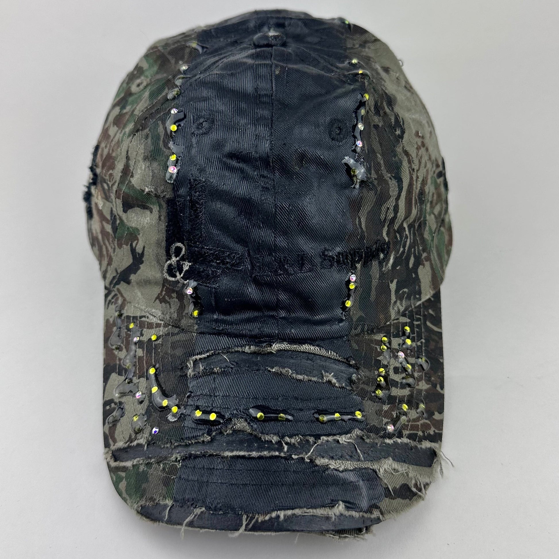 Front flat lay of ASTROBOI* L&L Supply green RealTree camo utility hat with purple and green rhinestones sealed in resin and black tattoo print detailing