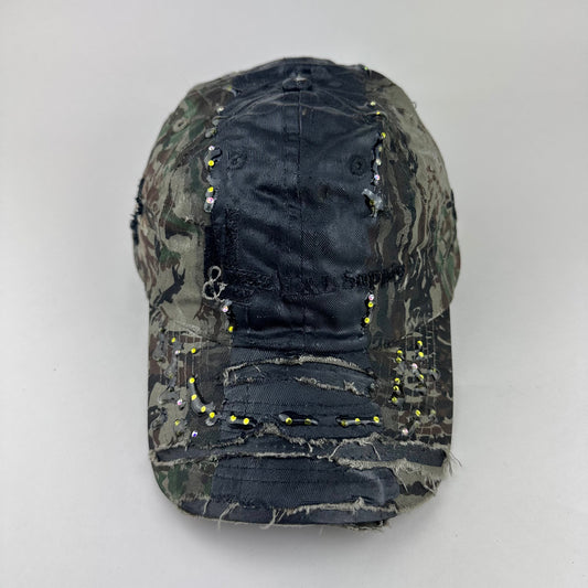 Front flat lay of ASTROBOI* L&L Supply green RealTree camo utility hat with purple and green rhinestones sealed in resin and black tattoo print detailing