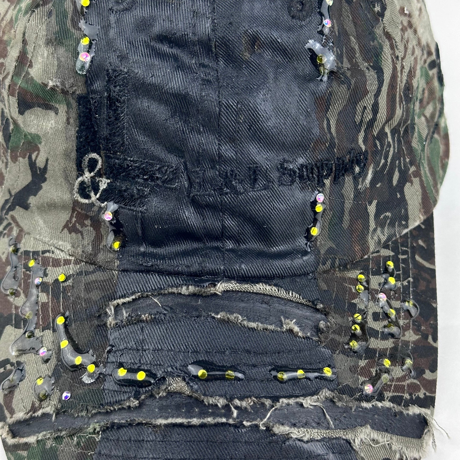 Flat front detail of ASTROBOI* L&L Supply green RealTree camo utility hat with purple and green rhinestones sealed in resin and black tattoo print detailing