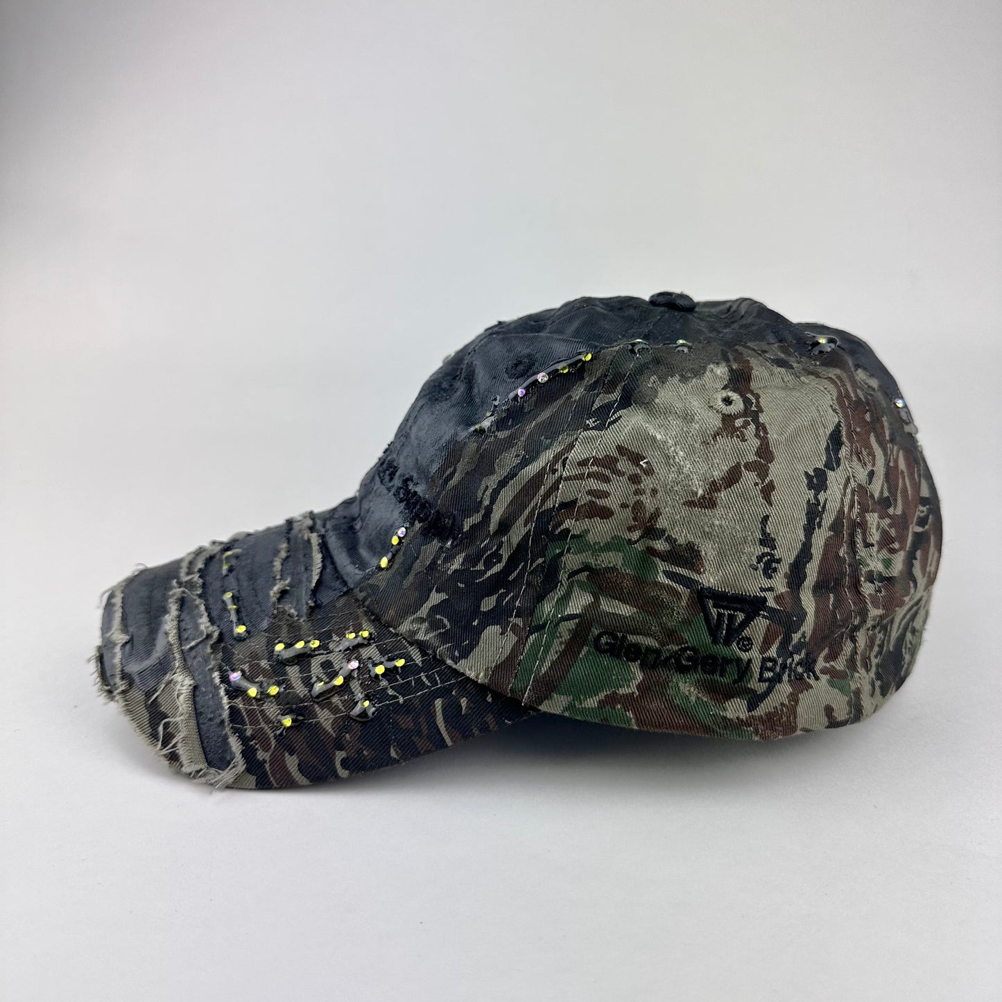 Flat lay left of ASTROBOI* L&L Supply green RealTree camo utility hat with purple and green rhinestones sealed in resin and black tattoo print detailing