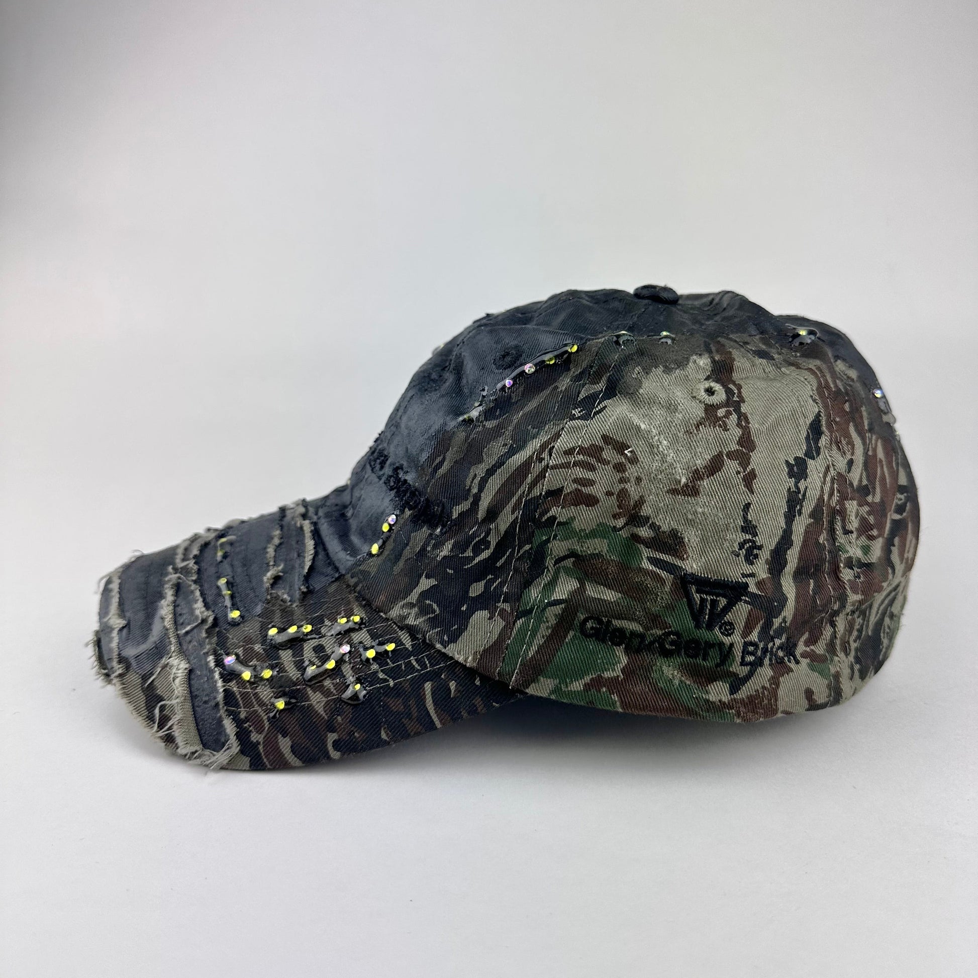 Flat lay left of ASTROBOI* L&L Supply green RealTree camo utility hat with purple and green rhinestones sealed in resin and black tattoo print detailing