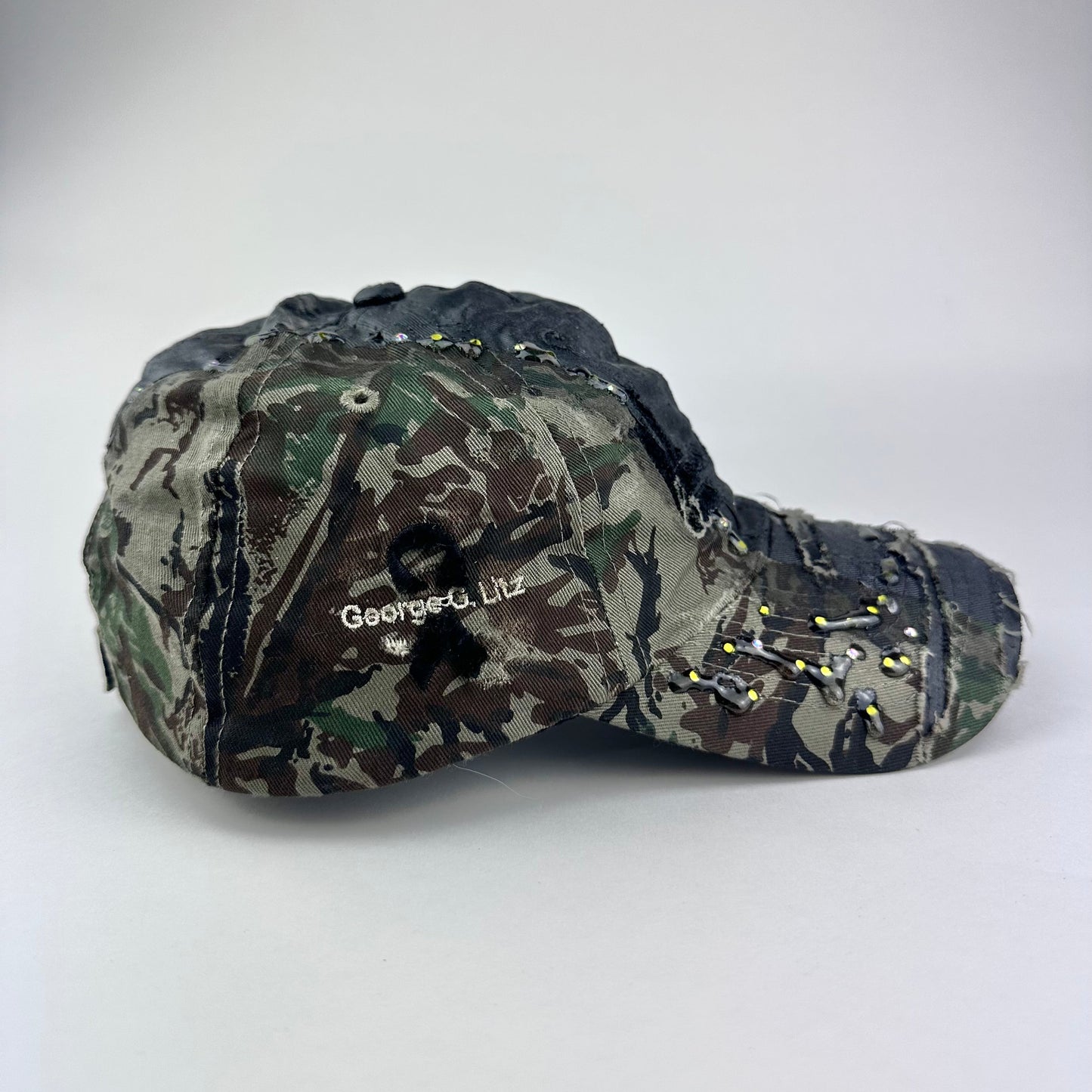 Flat lay right side of ASTROBOI* L&L Supply green RealTree camo utility hat with purple and green rhinestones sealed in resin and black tattoo print detailing