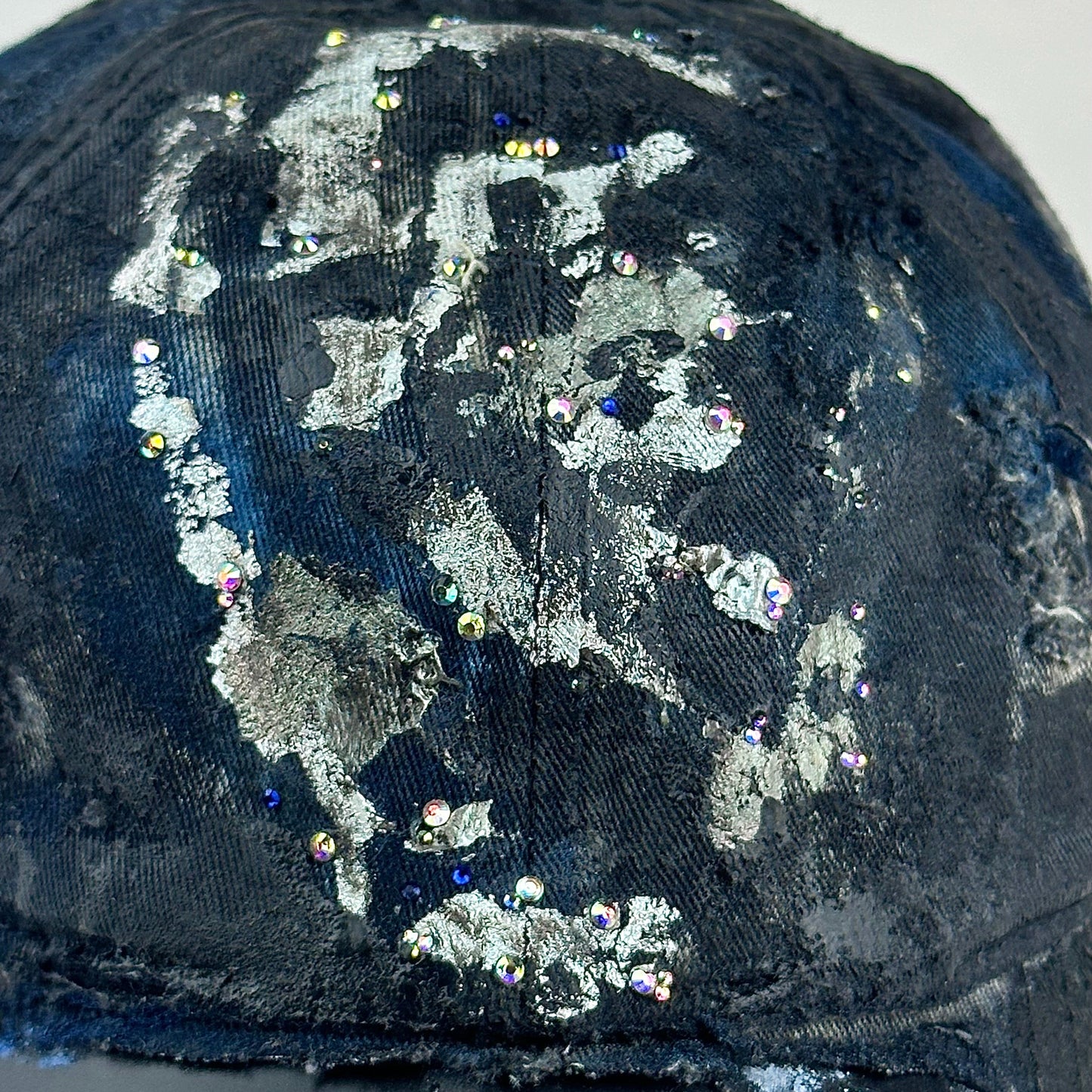 Close up detail on Black acid-washed denim hat with silver paint accents, multicolored rhinestones, and a ripped visor seam, customized by ASTROBOI* STUDIO.