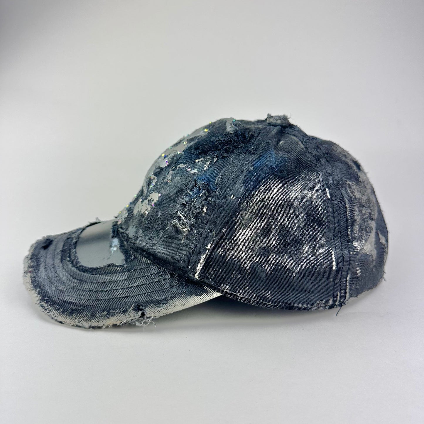 Flay lay left side on Black acid-washed denim hat with silver paint accents, multicolored rhinestones, and a ripped visor seam, customized by ASTROBOI* STUDIO.