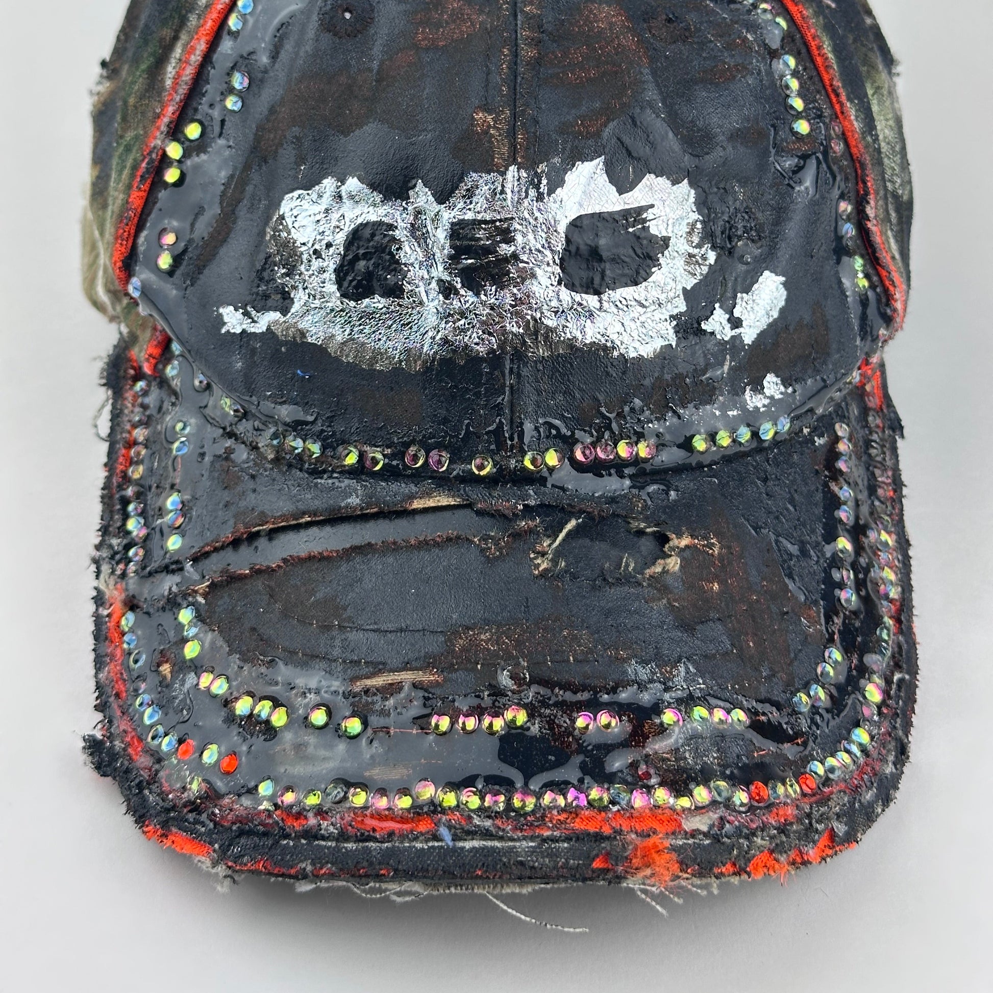 Flay front detail of of ASTROBOI* brown camouflage distressed hat with rhinestones sealed in clear resin and custom hand-painted detailing.