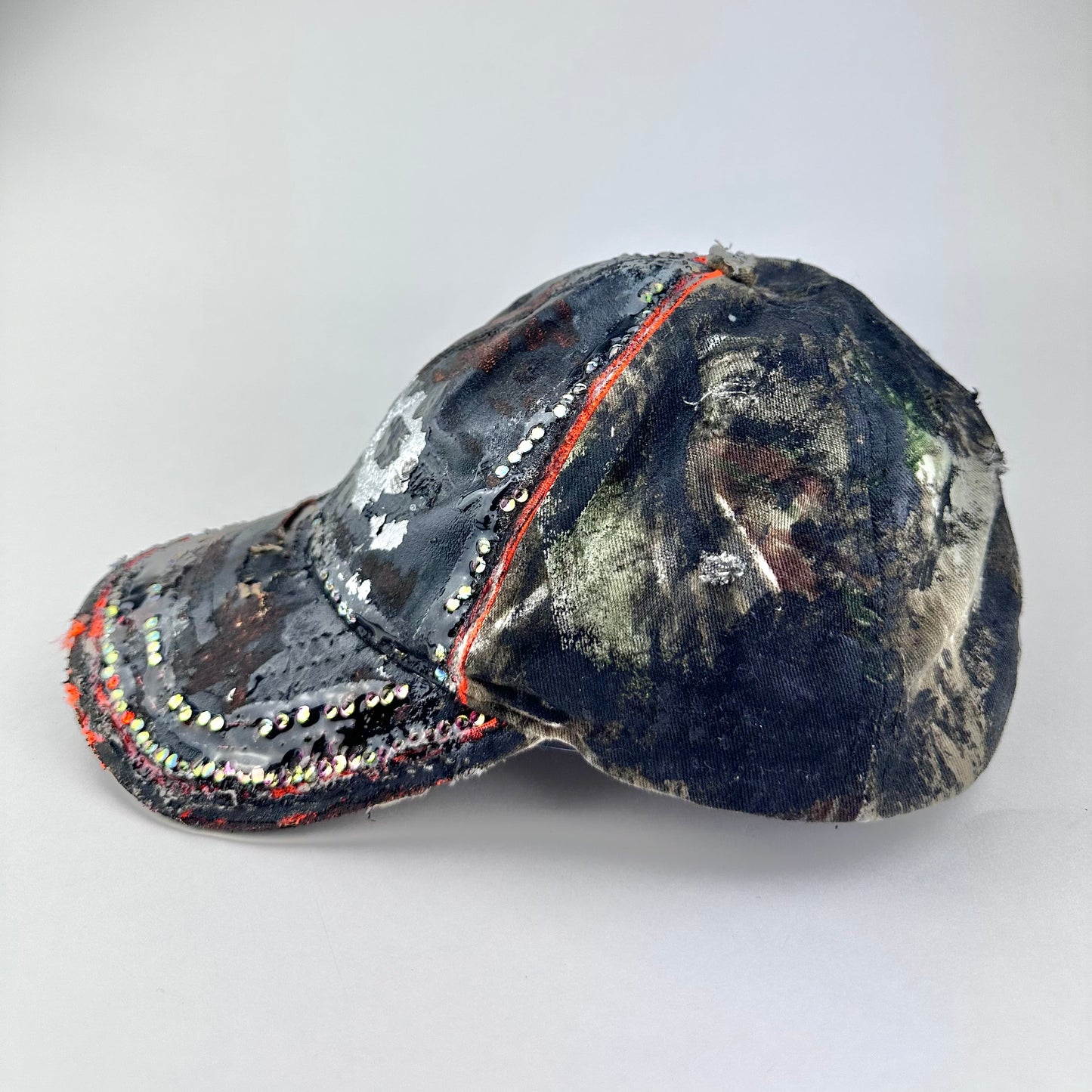 Flat front left of ASTROBOI* brown camouflage distressed hat with rhinestones sealed in clear resin and custom hand-painted detailing.