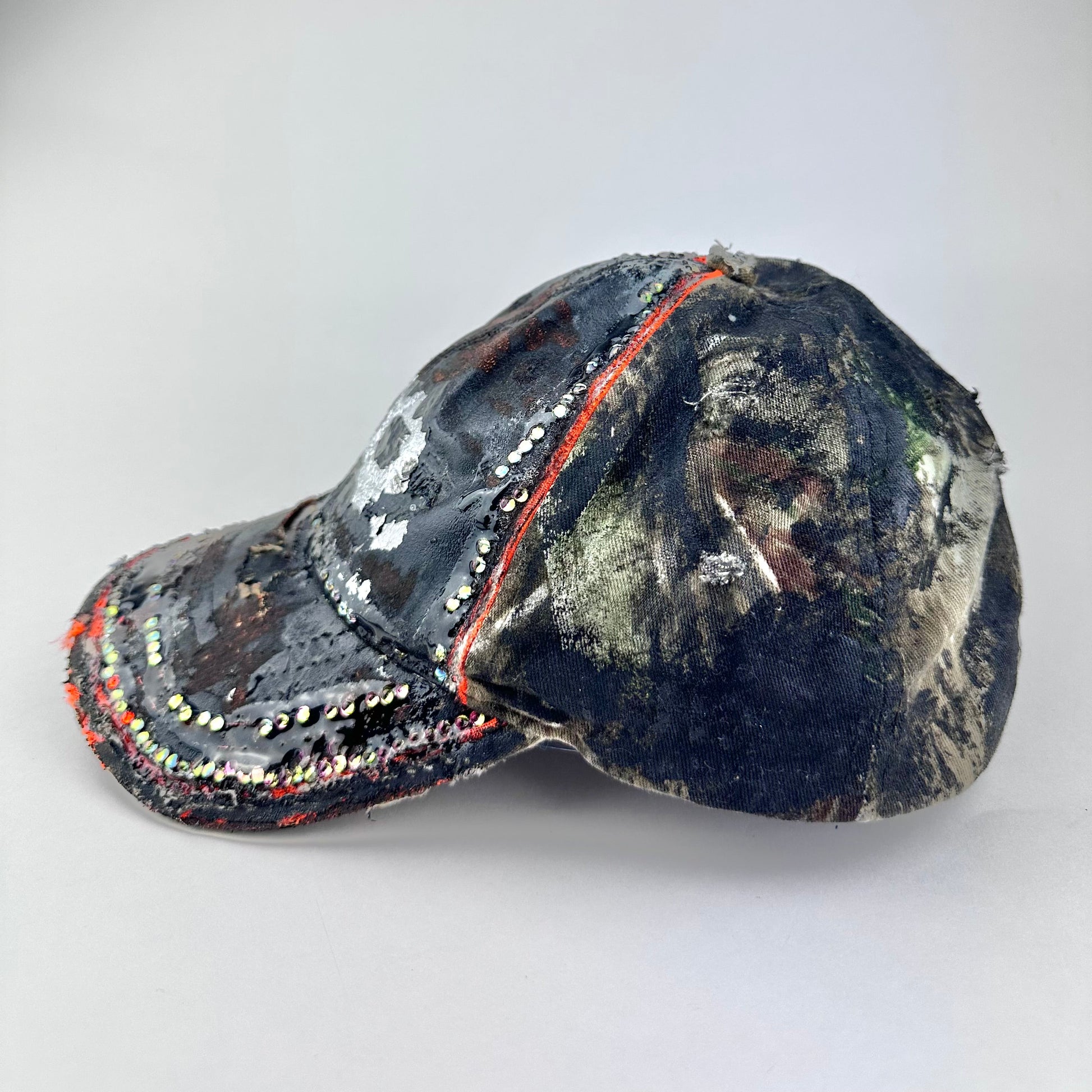 Flat front left of ASTROBOI* brown camouflage distressed hat with rhinestones sealed in clear resin and custom hand-painted detailing.
