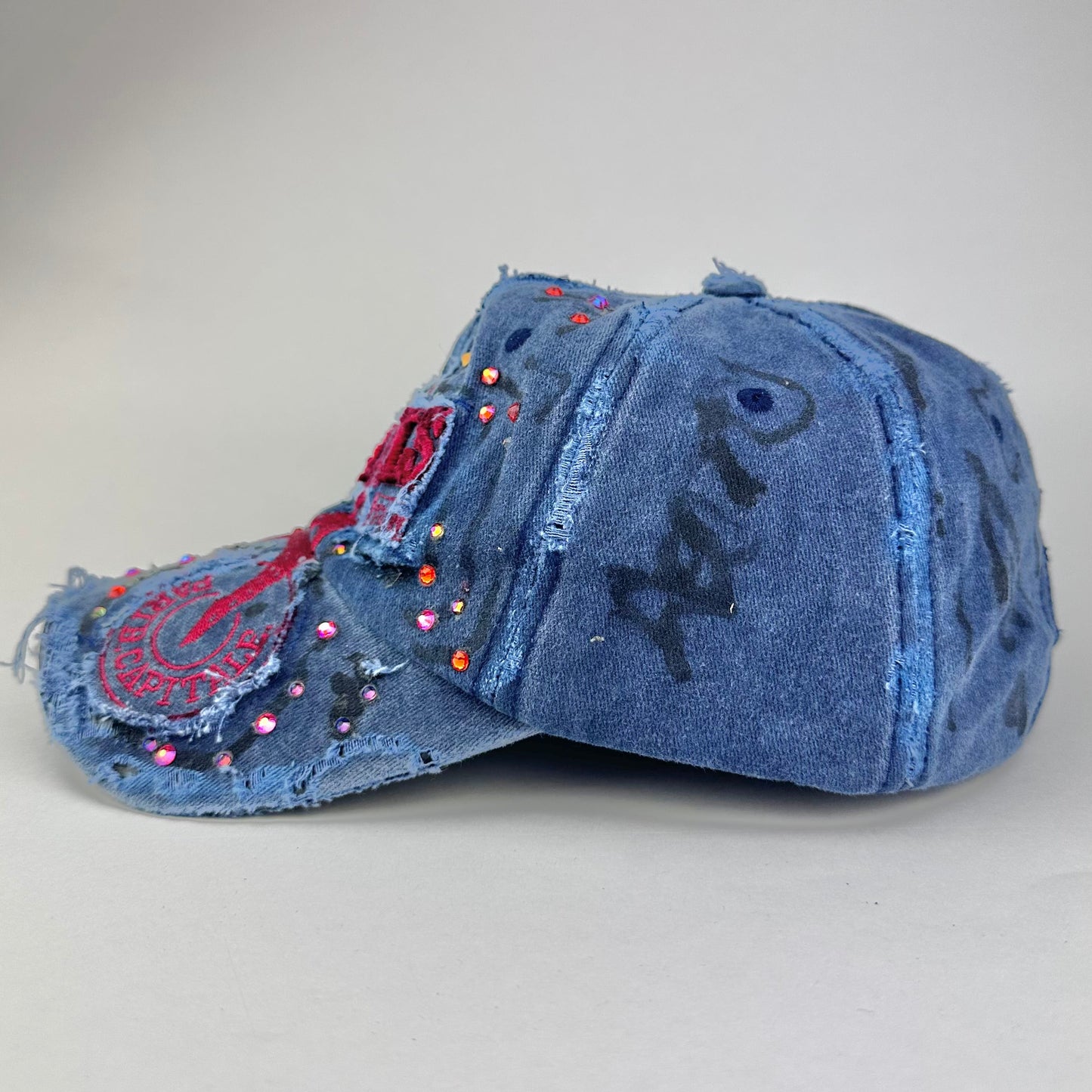 Left side of ASTROBOI* Lights of Paris blue baseball hat with Eiffel Tower embroidery, black graffiti print and red orange rhinestone crystals.