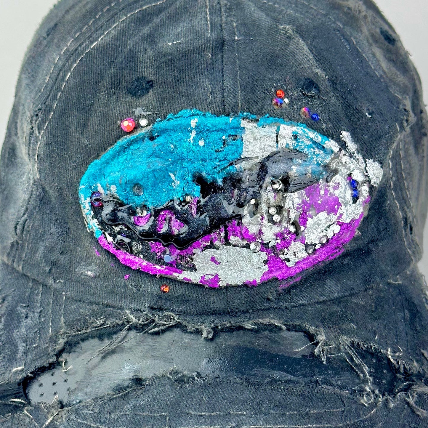 Logo detail on ASTROBOI* Technicolor black distressed hat with multicolor metallic New Orleans logo, rhinestones sealed in resin and ripped visor detail.