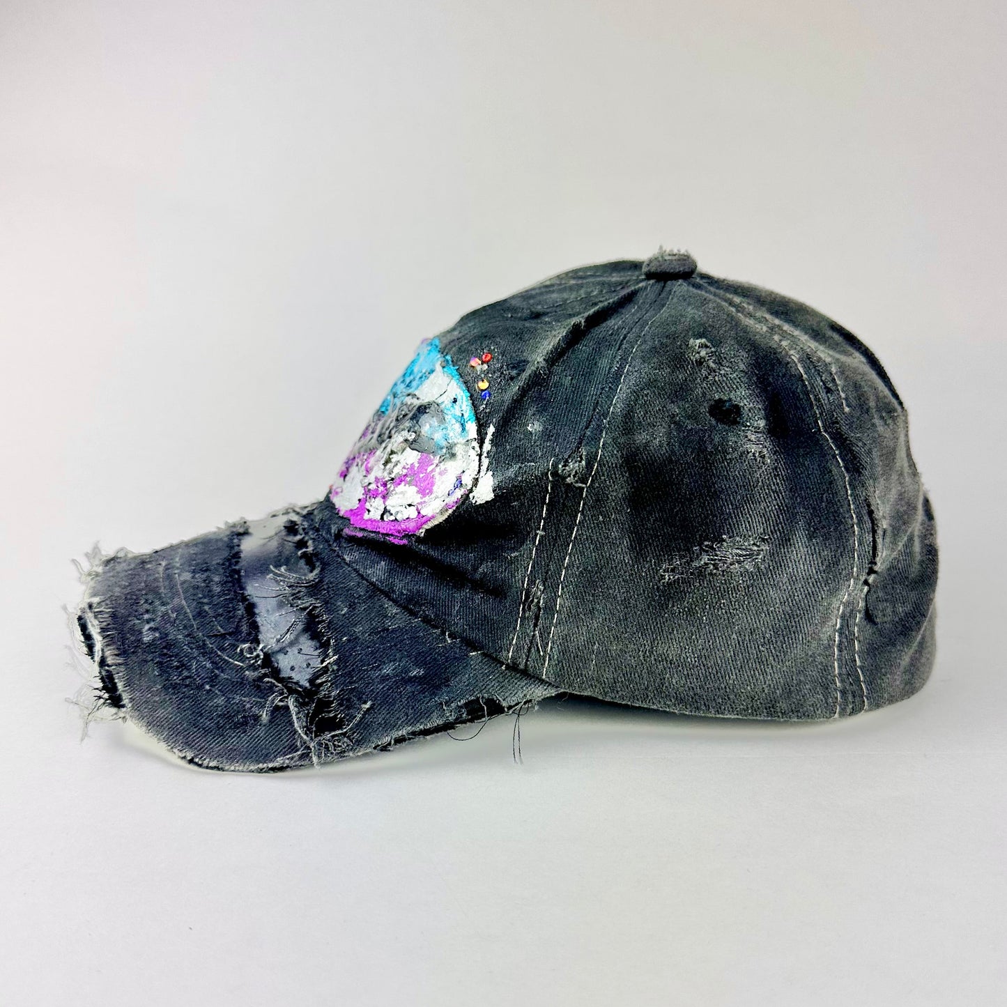 Left side of ASTROBOI* Technicolor black distressed hat with multicolor metallic New Orleans logo, rhinestones sealed in resin and ripped visor detail.