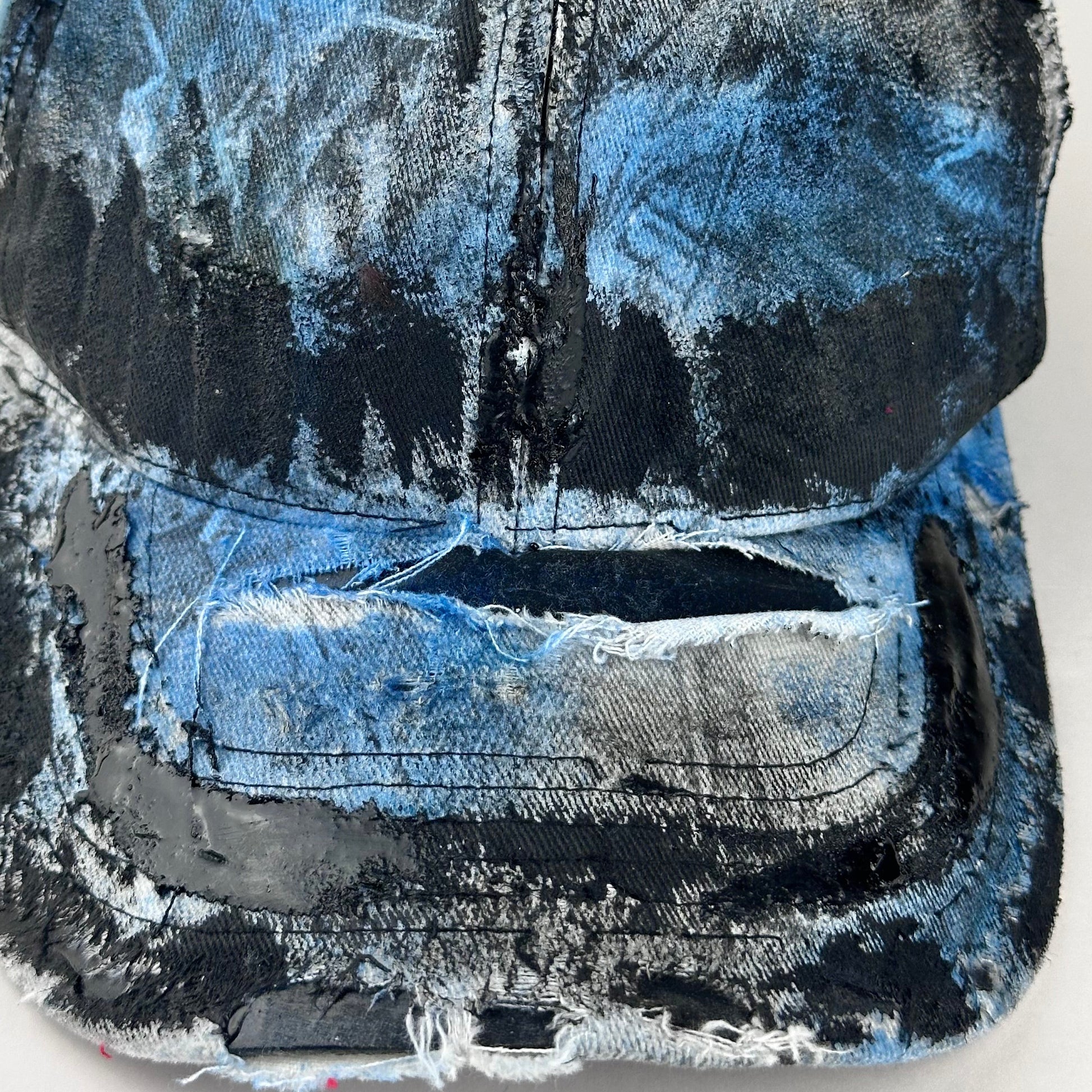 Close up detail of ASTROBOI* white trompe l’oeil denim print hat with blue and black paint, resin accents, and distressed detailing.