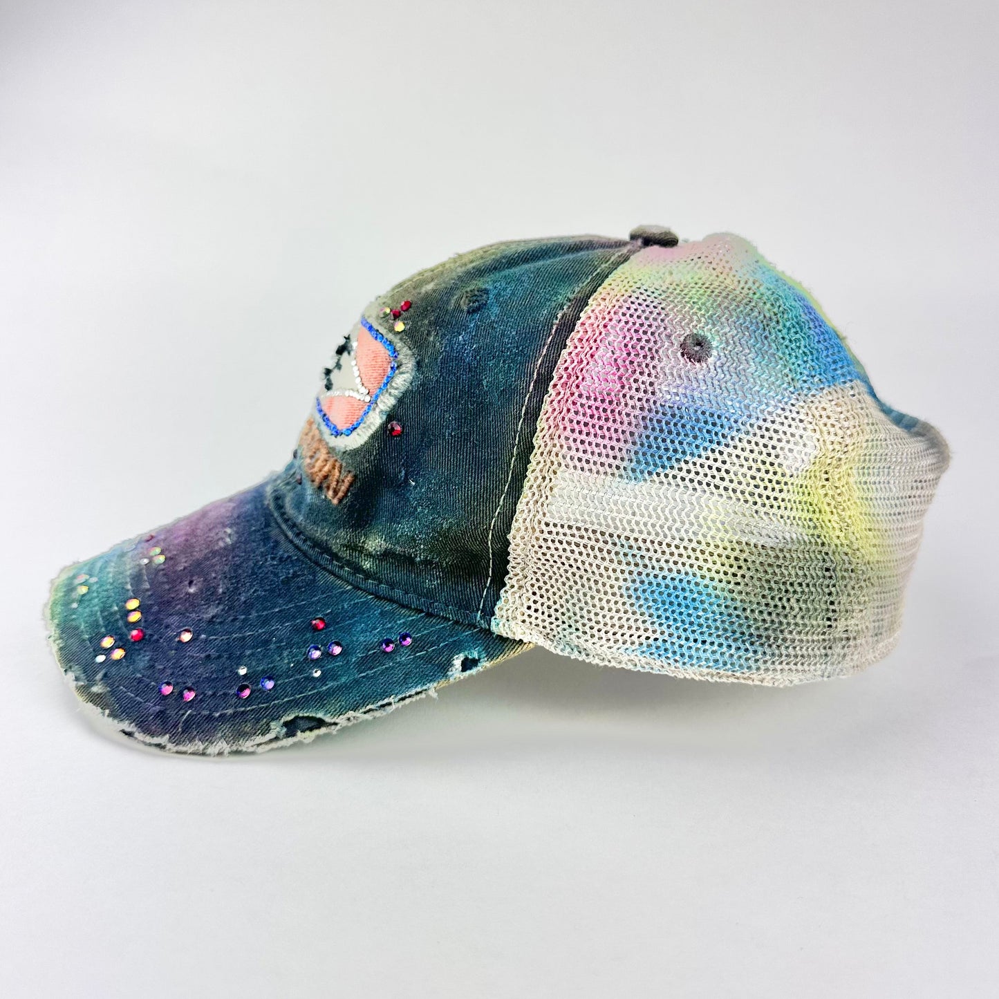 Left side of ASTROBOI* EASTON rainbow rhinestone mesh trucker hat with airbrushed confetti design, sparkling logo, and adjustable back strap.