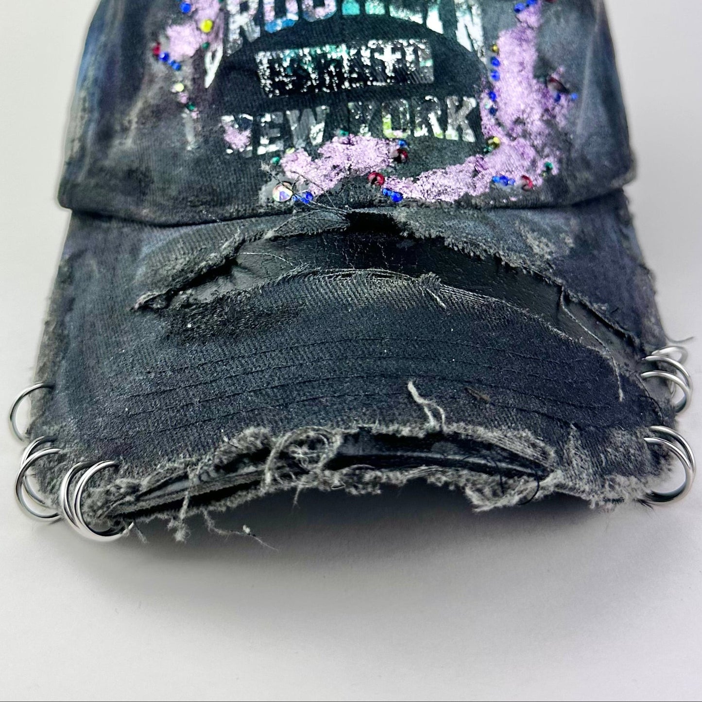 Visor detail on ASTROBOI* distressed black Brooklyn hat with metallic purple paint, multicolored rhinestones, green airbrush shading, and white front lettering.