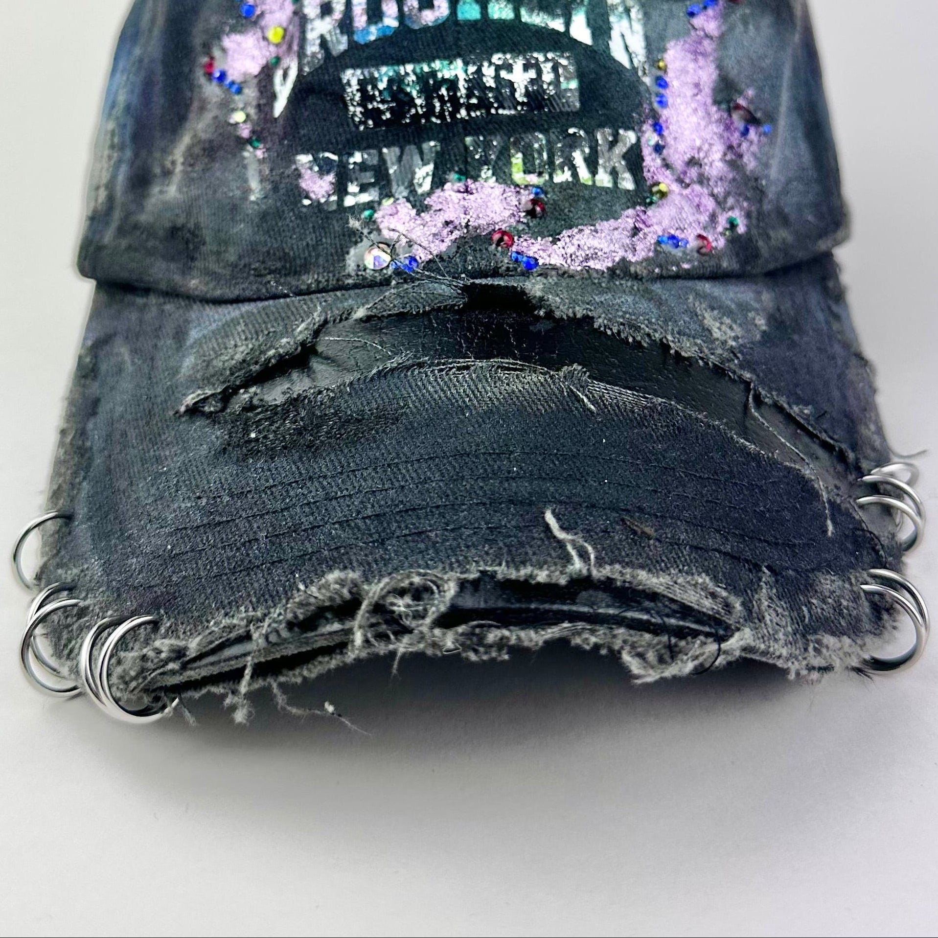 Visor detail on ASTROBOI* distressed black Brooklyn hat with metallic purple paint, multicolored rhinestones, green airbrush shading, and white front lettering.