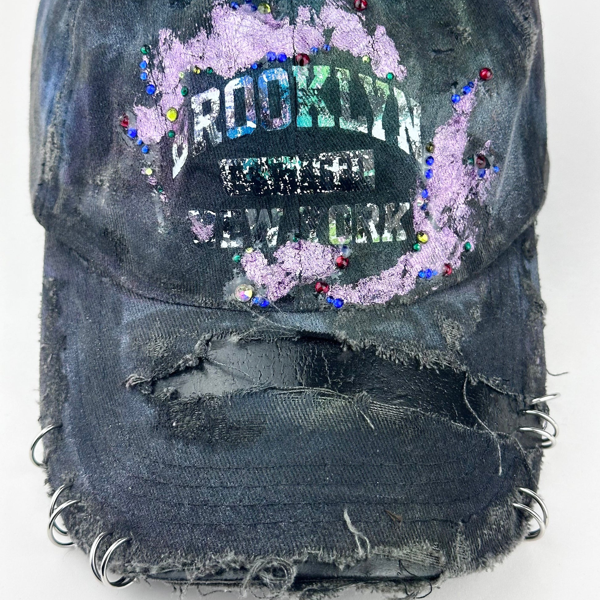 Close up of ASTROBOI* distressed black Brooklyn hat with metallic purple paint, multicolored rhinestones, green airbrush shading, and white front lettering.