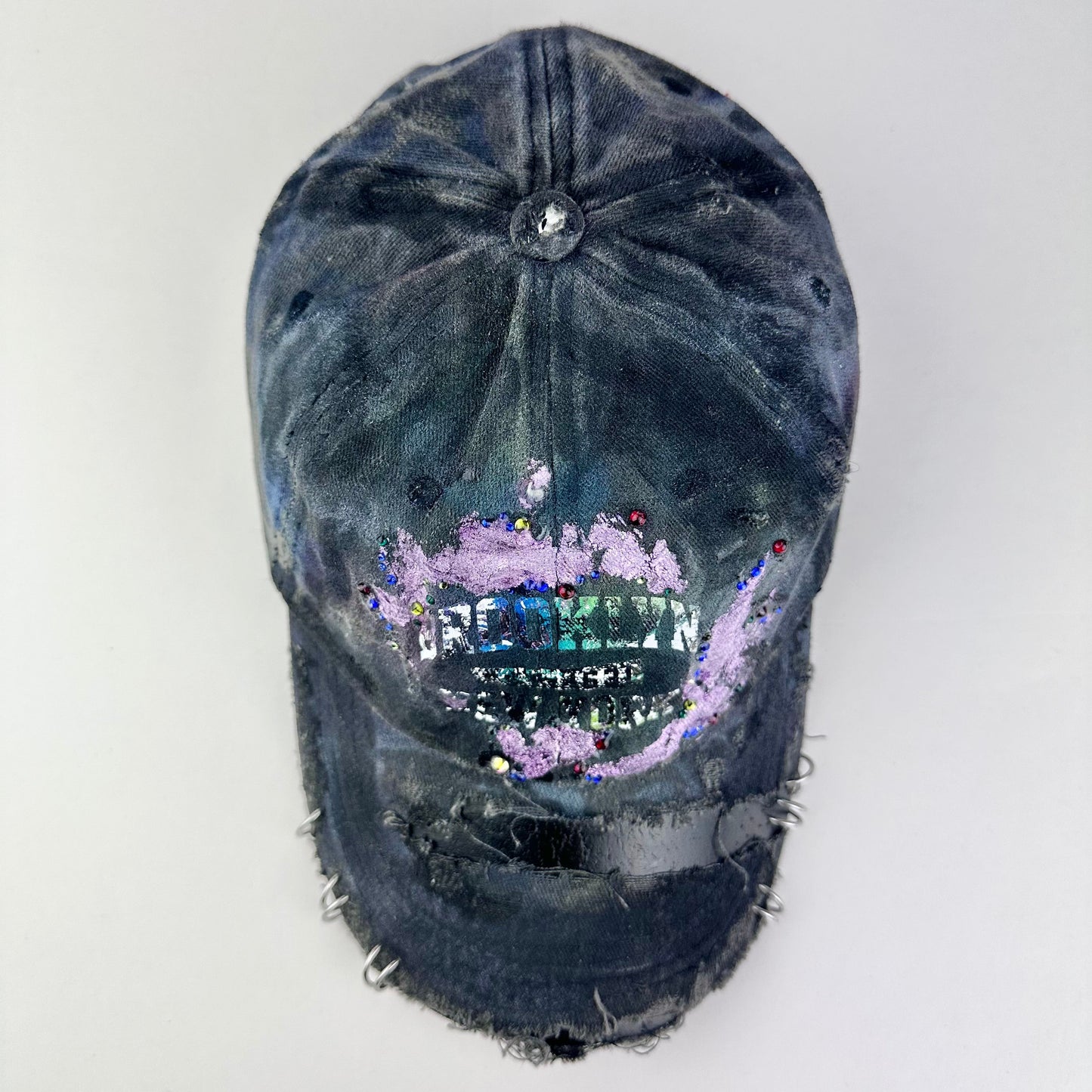Top of ASTROBOI* distressed black Brooklyn hat with metallic purple paint, multicolored rhinestones, green airbrush shading, and white front lettering.