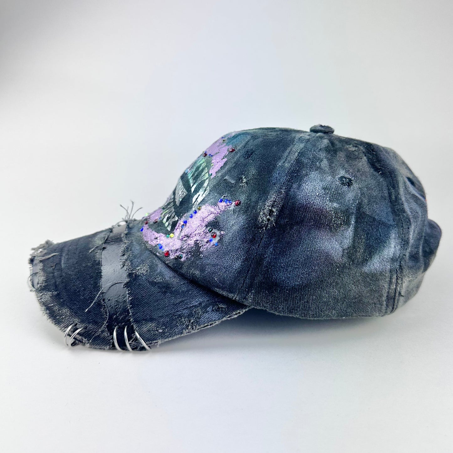 Left side of ASTROBOI* distressed black Brooklyn hat with metallic purple paint, multicolored rhinestones, green airbrush shading, and white front lettering.