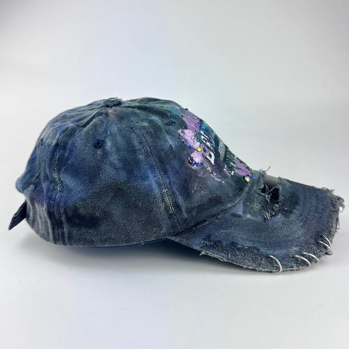 Right side of ASTROBOI* distressed black Brooklyn hat with metallic purple paint, multicolored rhinestones, green airbrush shading, and white front lettering.