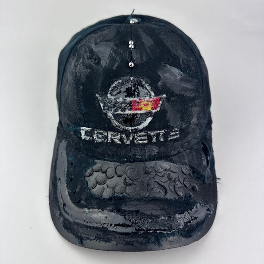 Cover image of “Black distressed ASTROBOI* x Corvette hat with resin-coated logo, stainless steel piercings, and deconstructed visor with circular peep holes.”