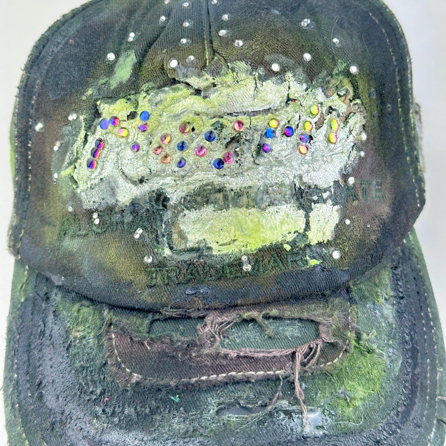 Close up of “Green and brown distressed ASTROBOI* Hawaii hat with rhinestones, metallic paint, and sterling-steel piercings, customized in NYC.”