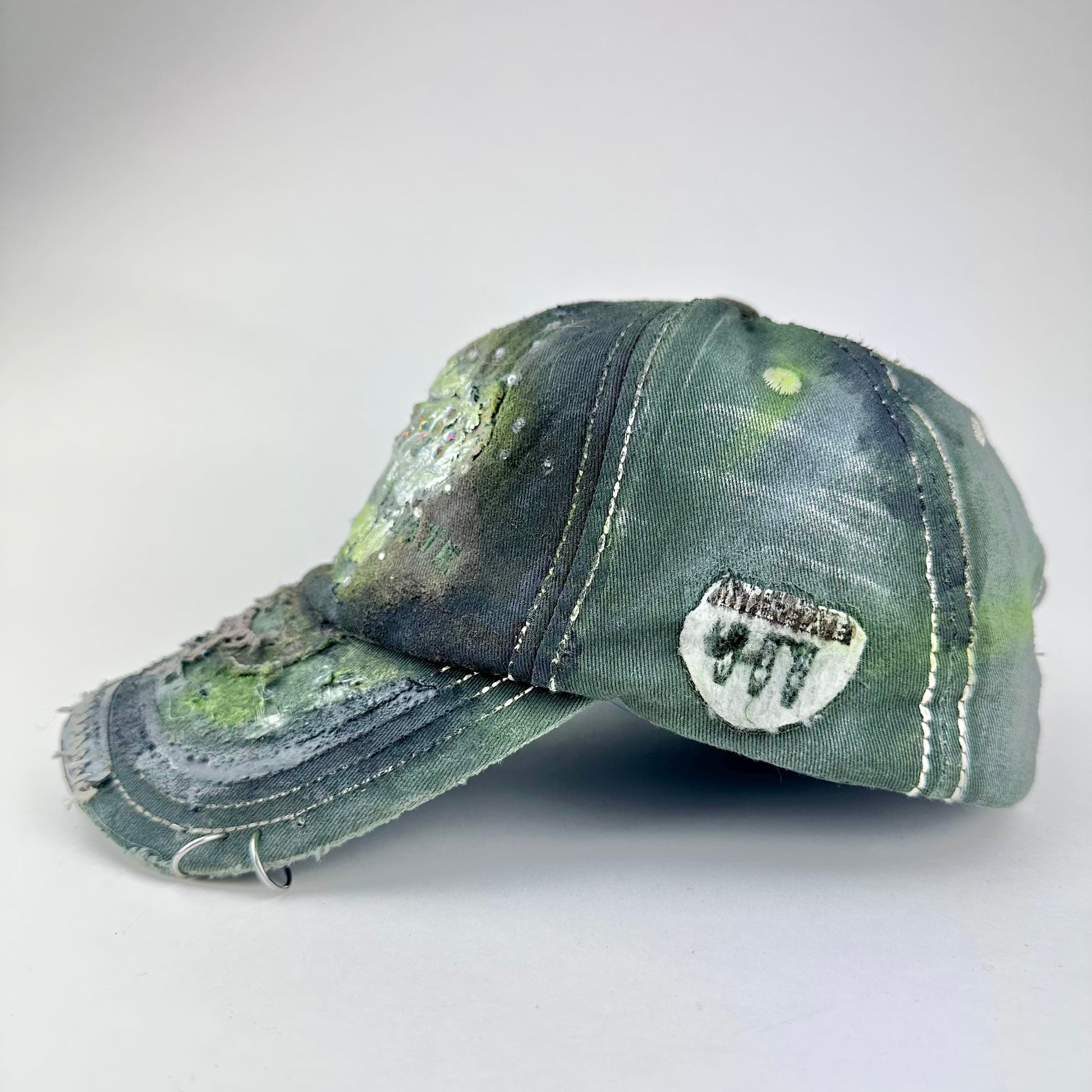 Left side of “Green and brown distressed ASTROBOI* Hawaii hat with rhinestones, metallic paint, and sterling-steel piercings, customized in NYC.”