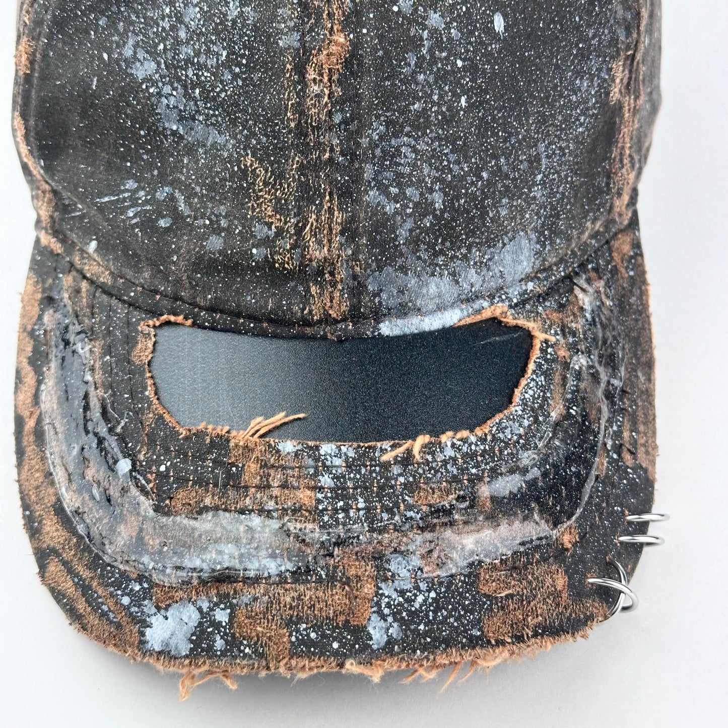 Close up of ASTROBOI* brown distressed leather hat with white paint splatter, stainless steel piercings on visor and glossy clear resin finish.