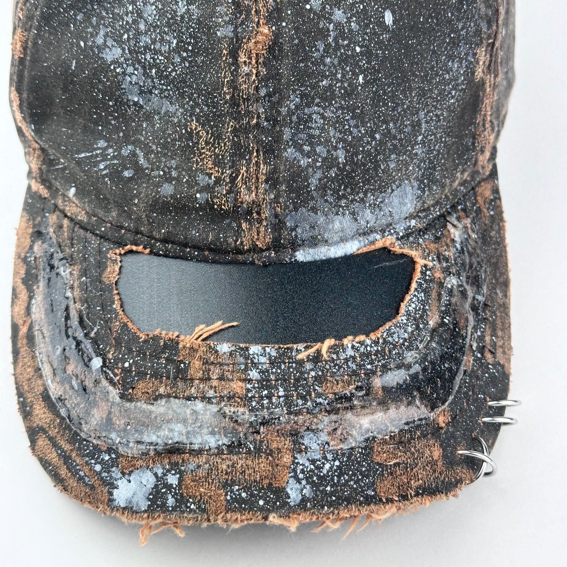 Close up of ASTROBOI* brown distressed leather hat with white paint splatter, stainless steel piercings on visor and glossy clear resin finish.