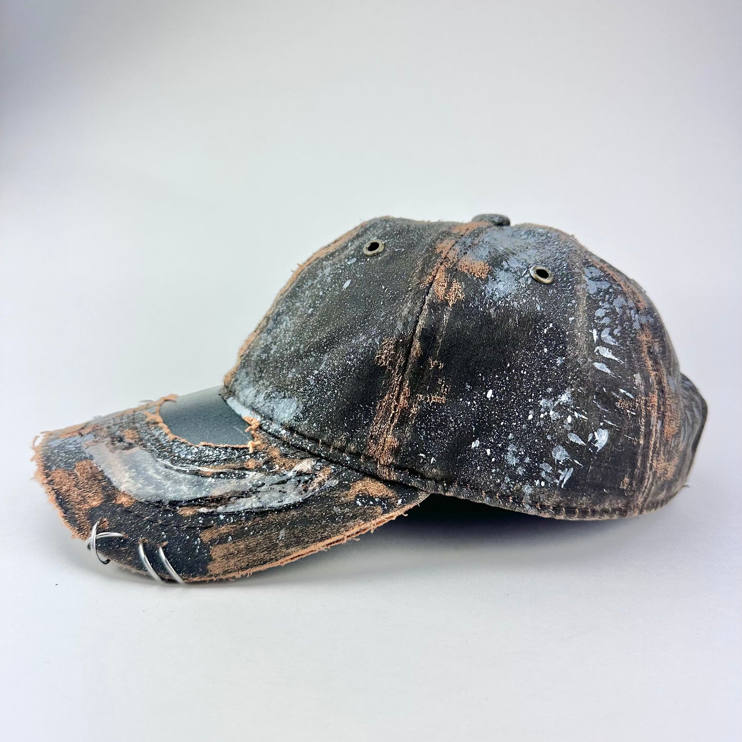 Left of ASTROBOI* brown distressed leather hat with white paint splatter, stainless steel piercings on visor and glossy clear resin finish.