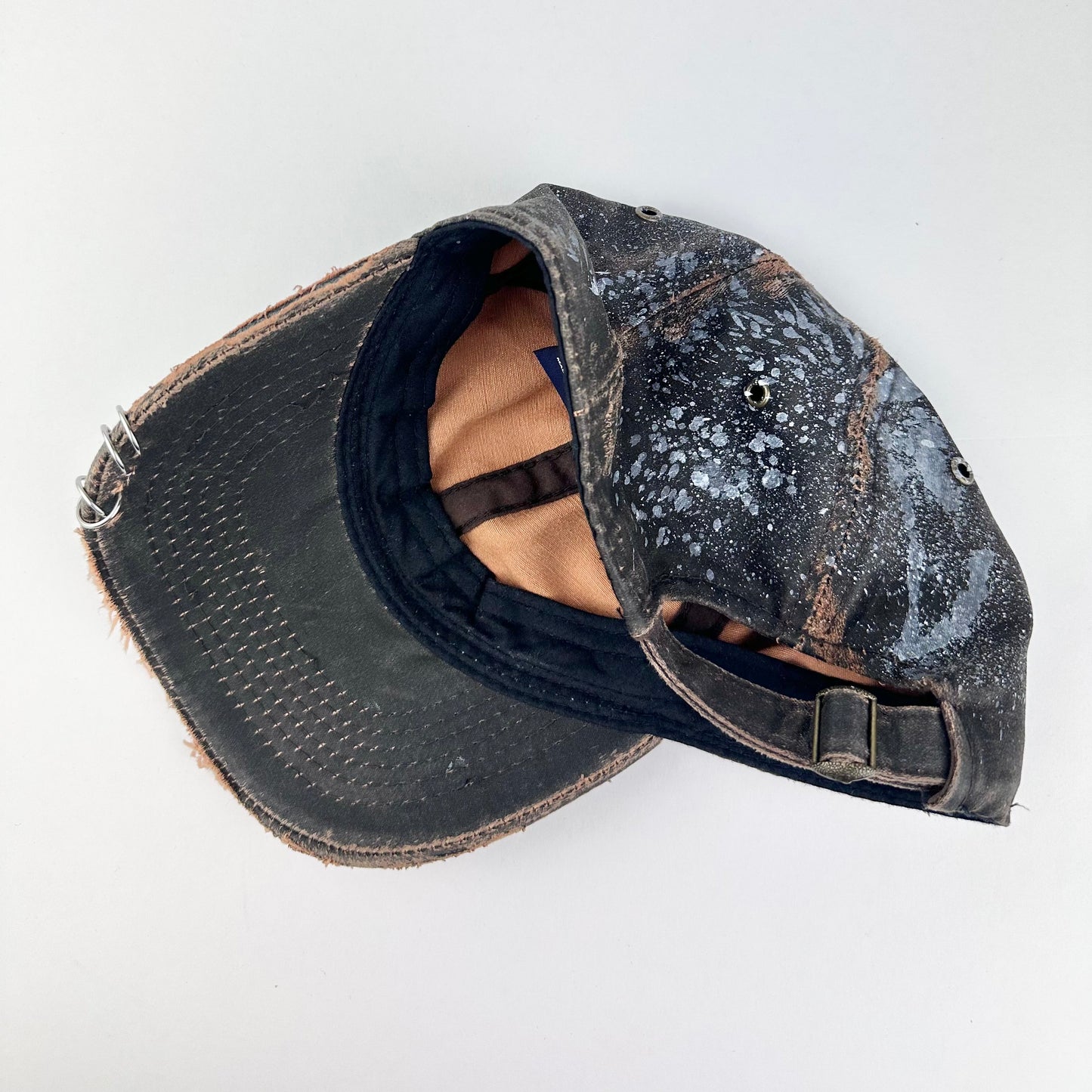 Back side of ASTROBOI* brown distressed leather hat with white paint splatter, stainless steel piercings on visor and glossy clear resin finish.