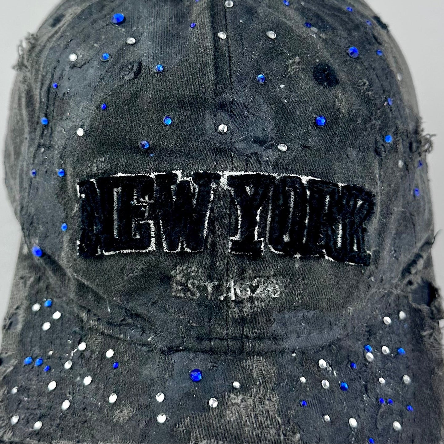 Close up of ASTROBOI* 'Oceane' black distressed half visor hat featuring New York logo, rhinestone accents, black metallic paint, and raw hem edge.