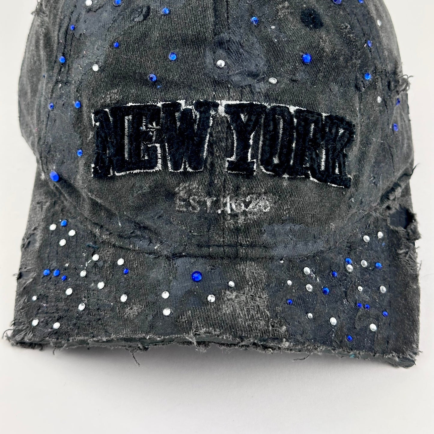 Visor detail on ASTROBOI* 'Oceane' black distressed half visor hat featuring New York logo, rhinestone accents, black metallic paint, and raw hem edge.