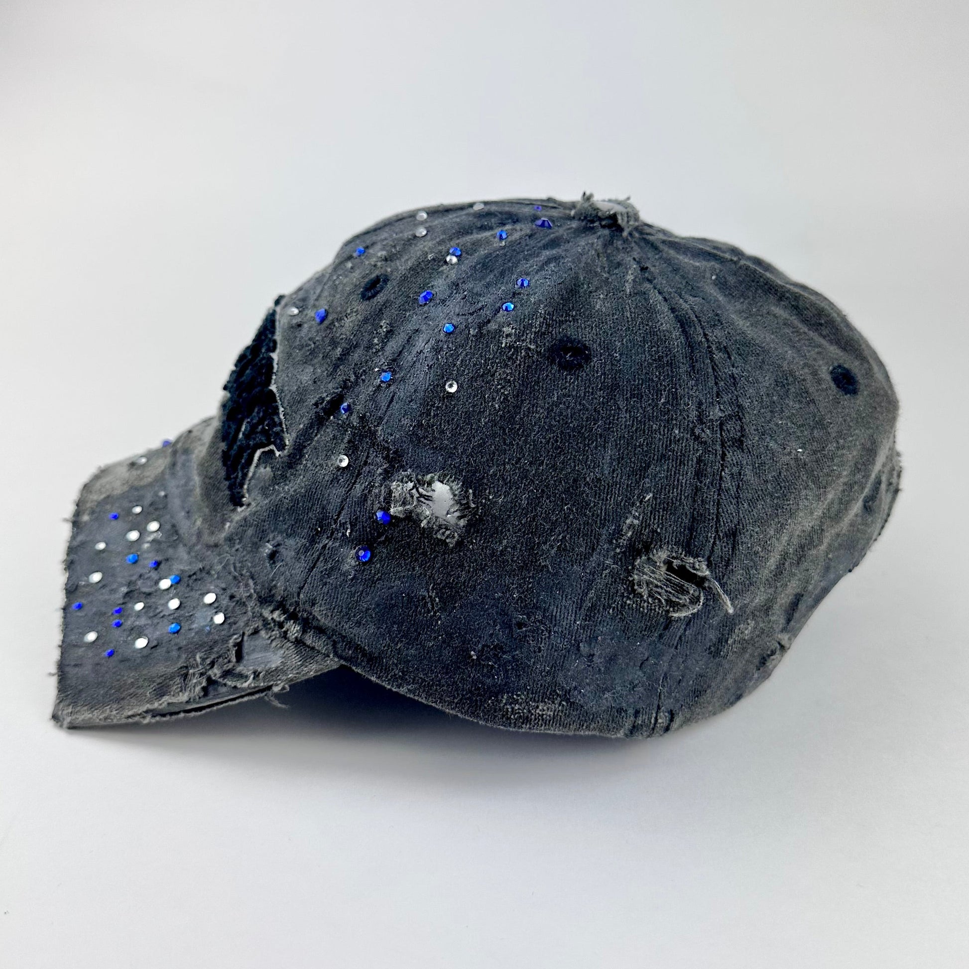 Left side of ASTROBOI* 'Oceane' black distressed half visor hat featuring New York logo, rhinestone accents, black metallic paint, and raw hem edge.