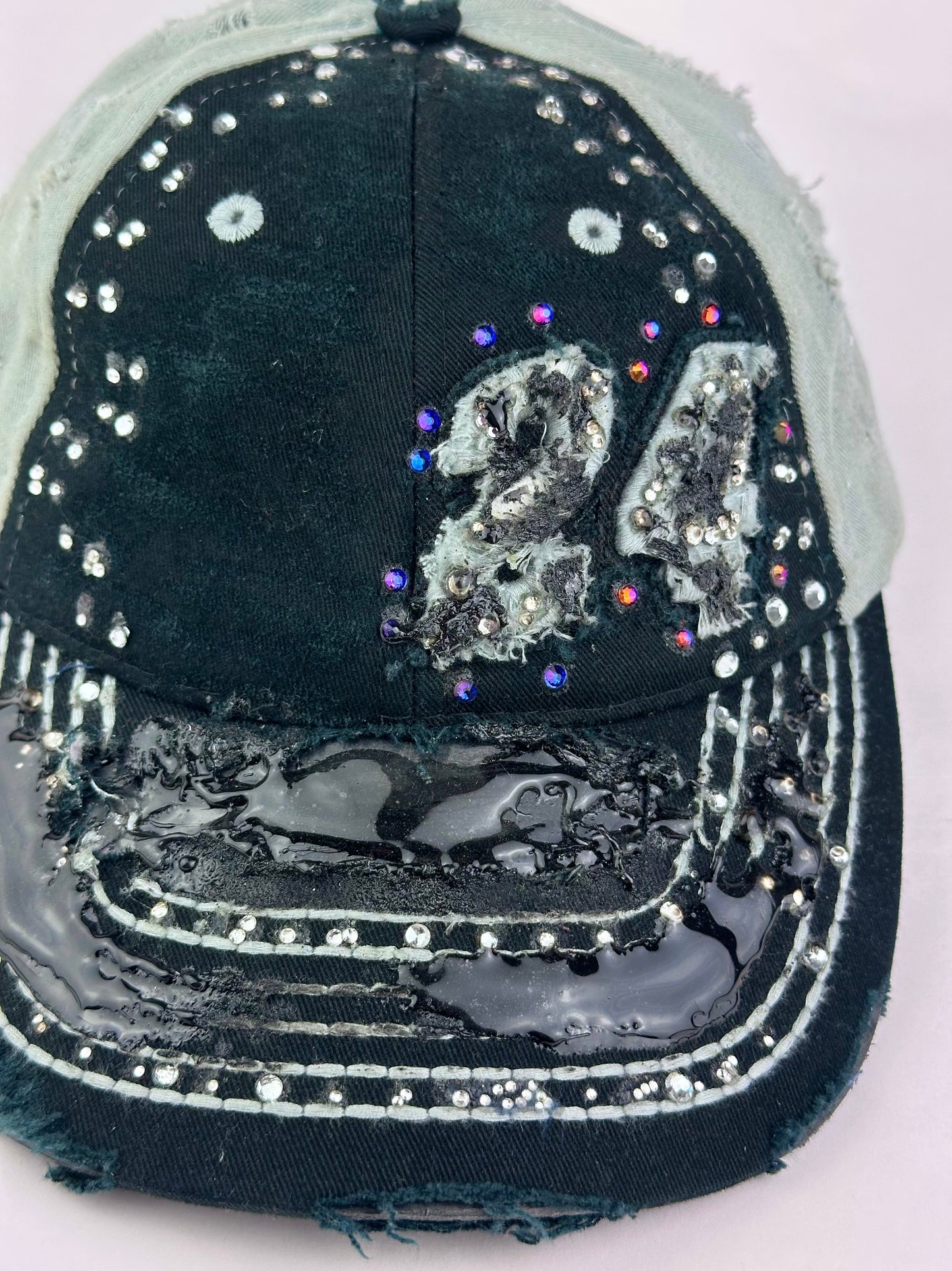 Close up detail of ASTROBOI* “24” black racing cap with silver rhinestones, white stitching, glossy black resin details, and adjustable Velcro back closure.