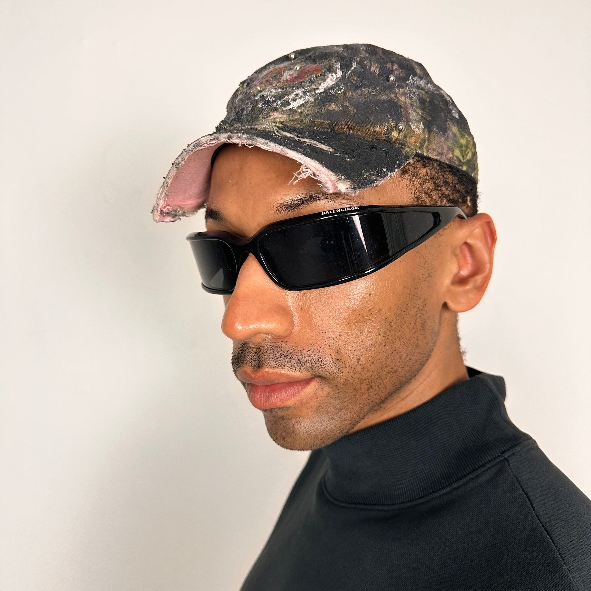 Front left side of ASTROBOI* x Mossy Oak brown real tree camo half visor hat with rhinestone resin logo and stainless steel piercings.