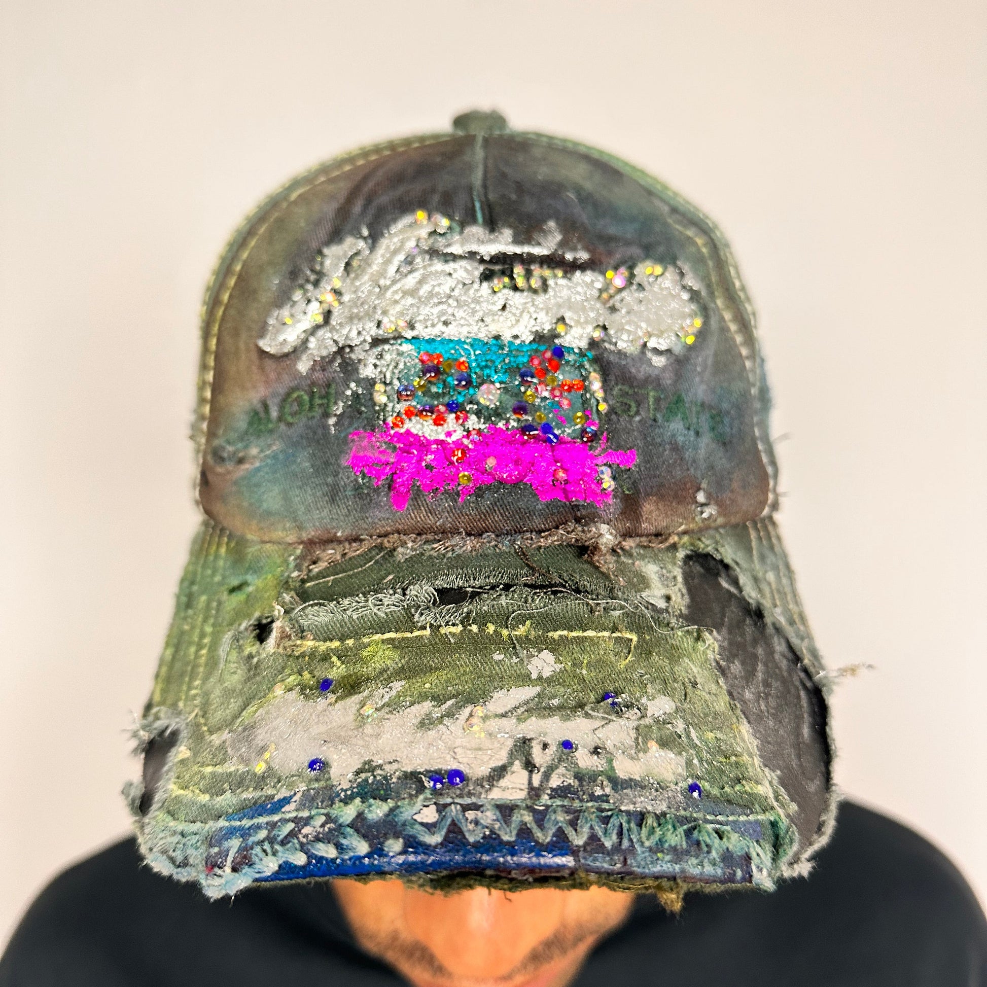 On body front shot of Brown-green ASTROBOI* hat featuring a metallic Hawaii logo with multicolored rhinestones, distressed visor, and resin-coated safety pin detail.