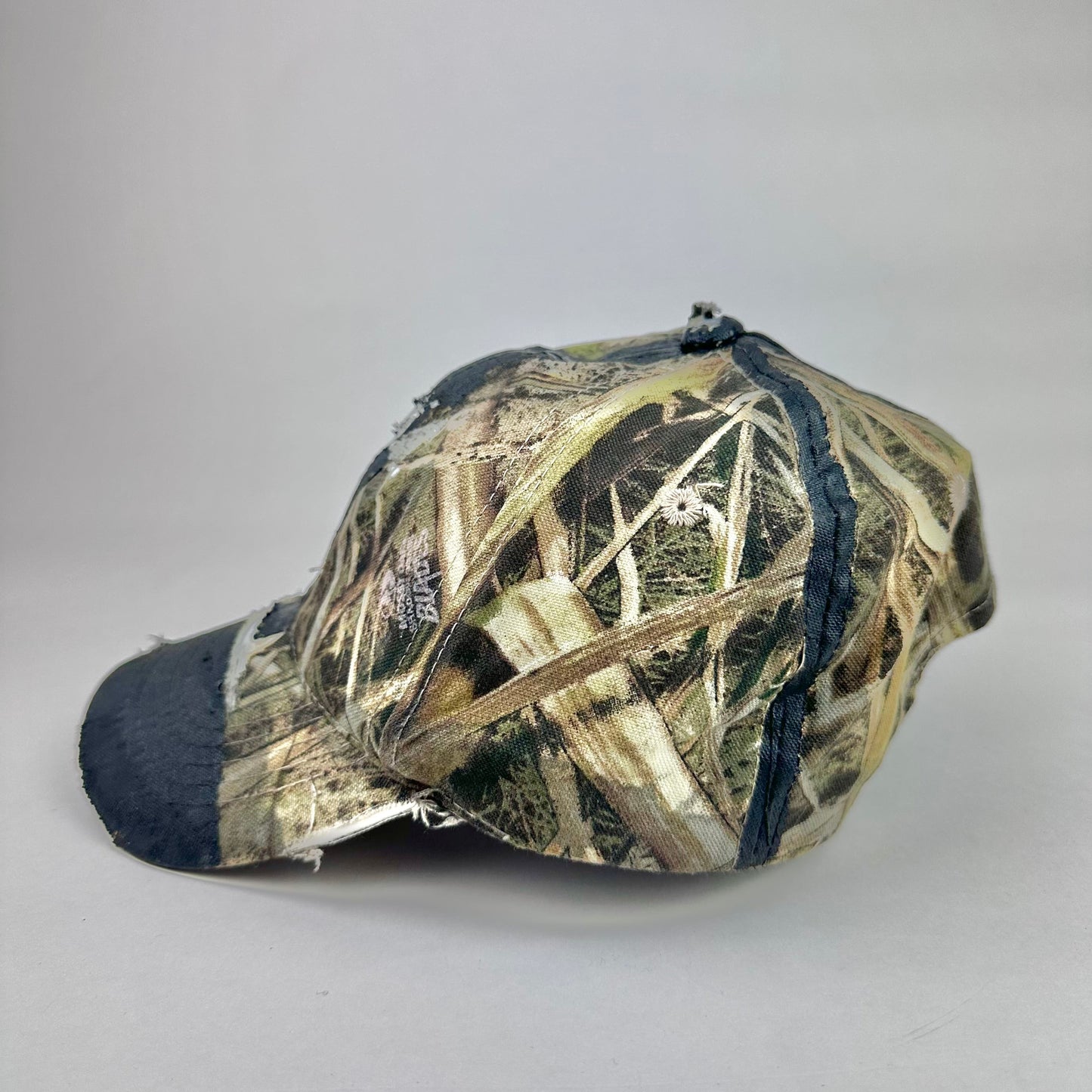 Left side of ASTROBOI* x Mossy Oak brown RealTree camouflage tattoo hat with SWA embroidery, black painted distressing, youth size, custom NYC piece
