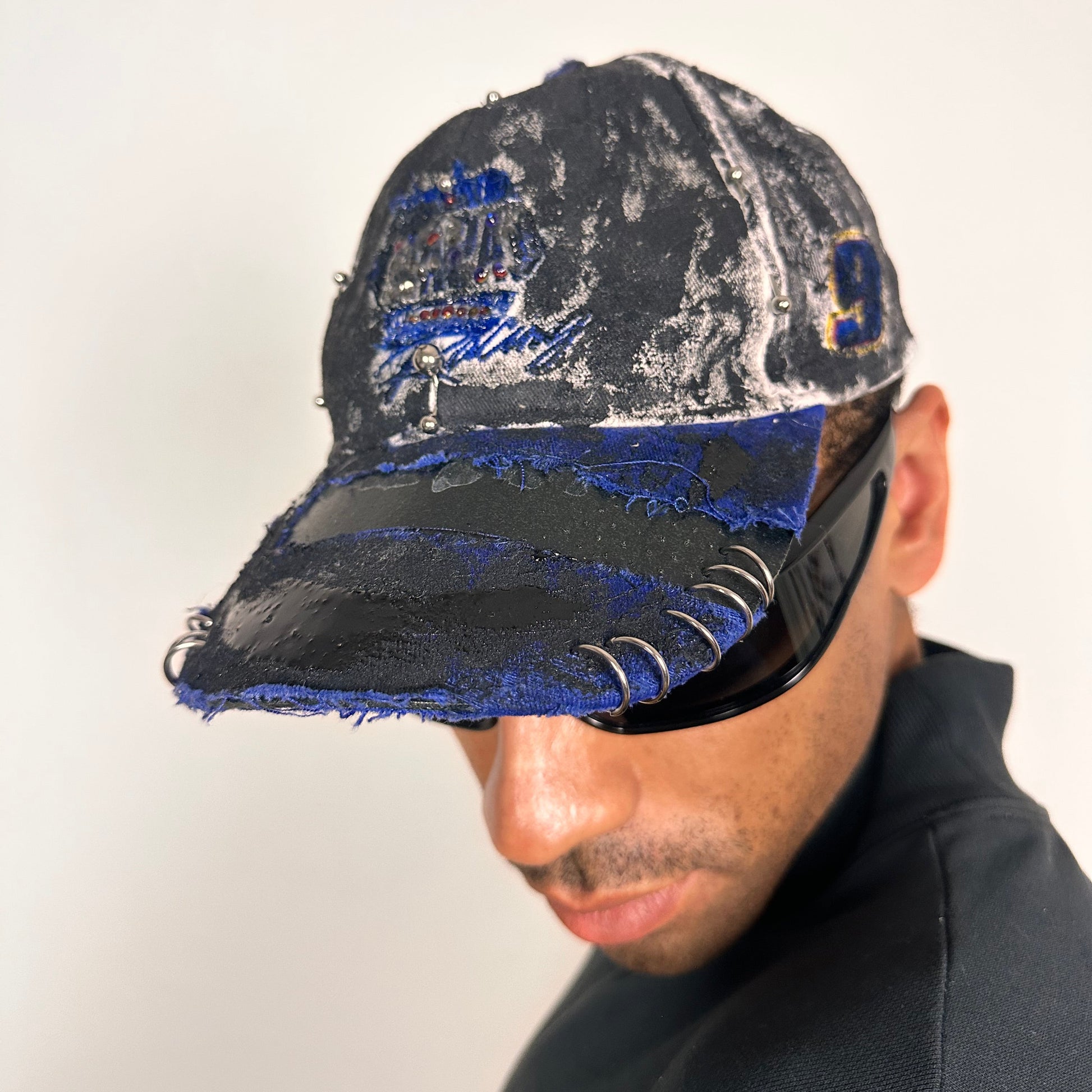 Front left side of Blue and white ASTROBOI* NAPA hat with rhinestone-covered logo sealed in clear resin, stainless steel piercings, and a deconstructed visor, handcrafted in NYC.