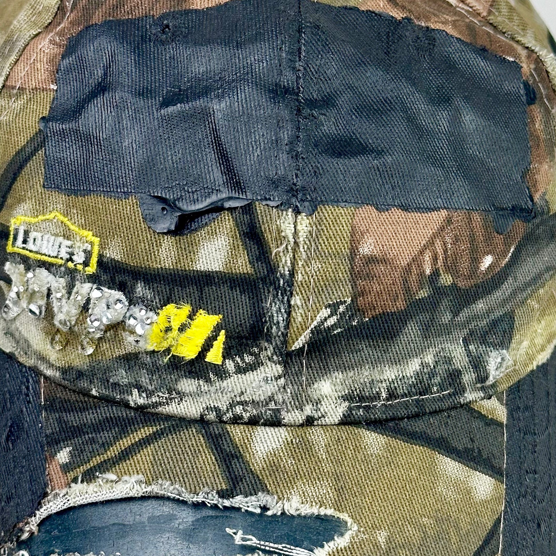 Still life front detail of ASTROBOI* x Lowe’s MVP brown RealTree camouflage hat with resin-sealed rhinestone logo, black waxed tattoo print, adjustable strap, custom NYC piece